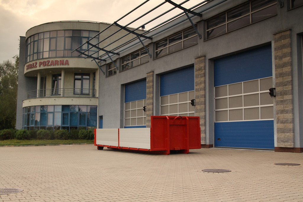 
Na pierwszym planie widoczna jest czerwona platforma ze srebrnymi burtami służąca do transportu sprzętu. W tle widoczny jest szary budynek z niebieskimi bramami Jednostki Ratowniczo-Gaśniczej numer 17 z dużym napisem Straż Pożarna.
