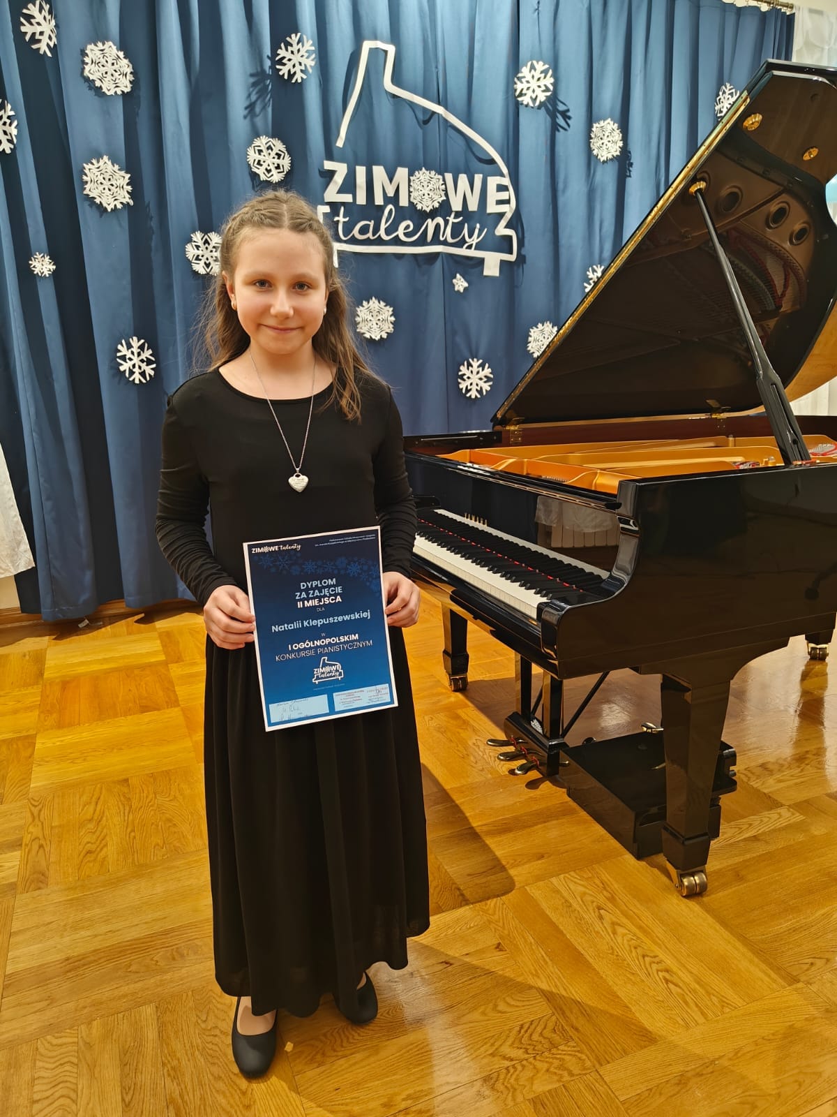Na zdjęciu znajduje się uśmiechnięta dziewczynka w czarnej długiej sukience , w rękach trzyma dyplom konkursowy. W tle znajduje się niebieska kotara z zimową dekoracją oraz czarny fortepian.