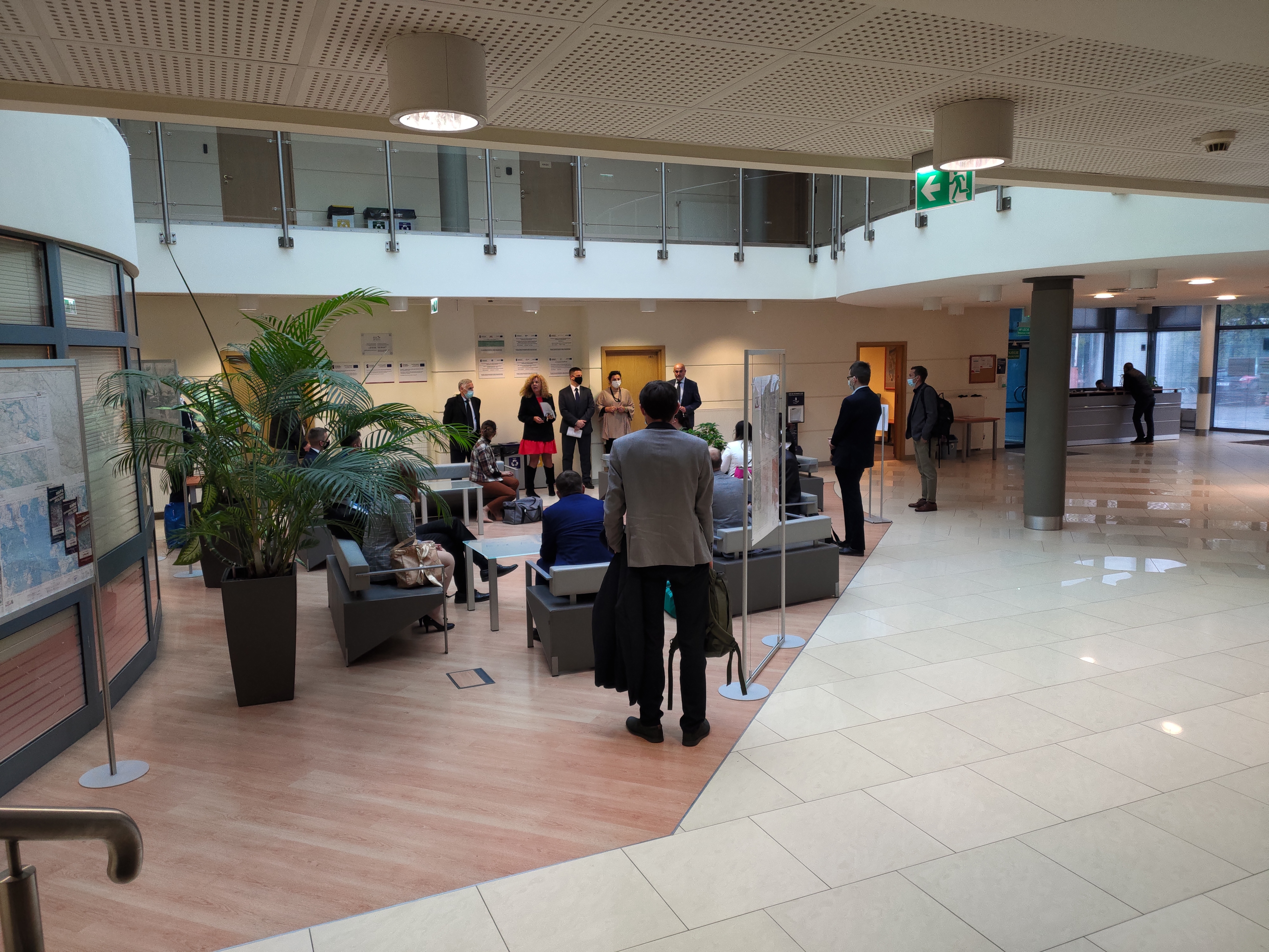 Zdjęcie przedstawia hol w siedzibie GUGiK przy ul. Olbrachta 94B z osobami przystępującymi do egzaminu słuchającymi wyjaśnień członków Komisji Egzaminacyjnej