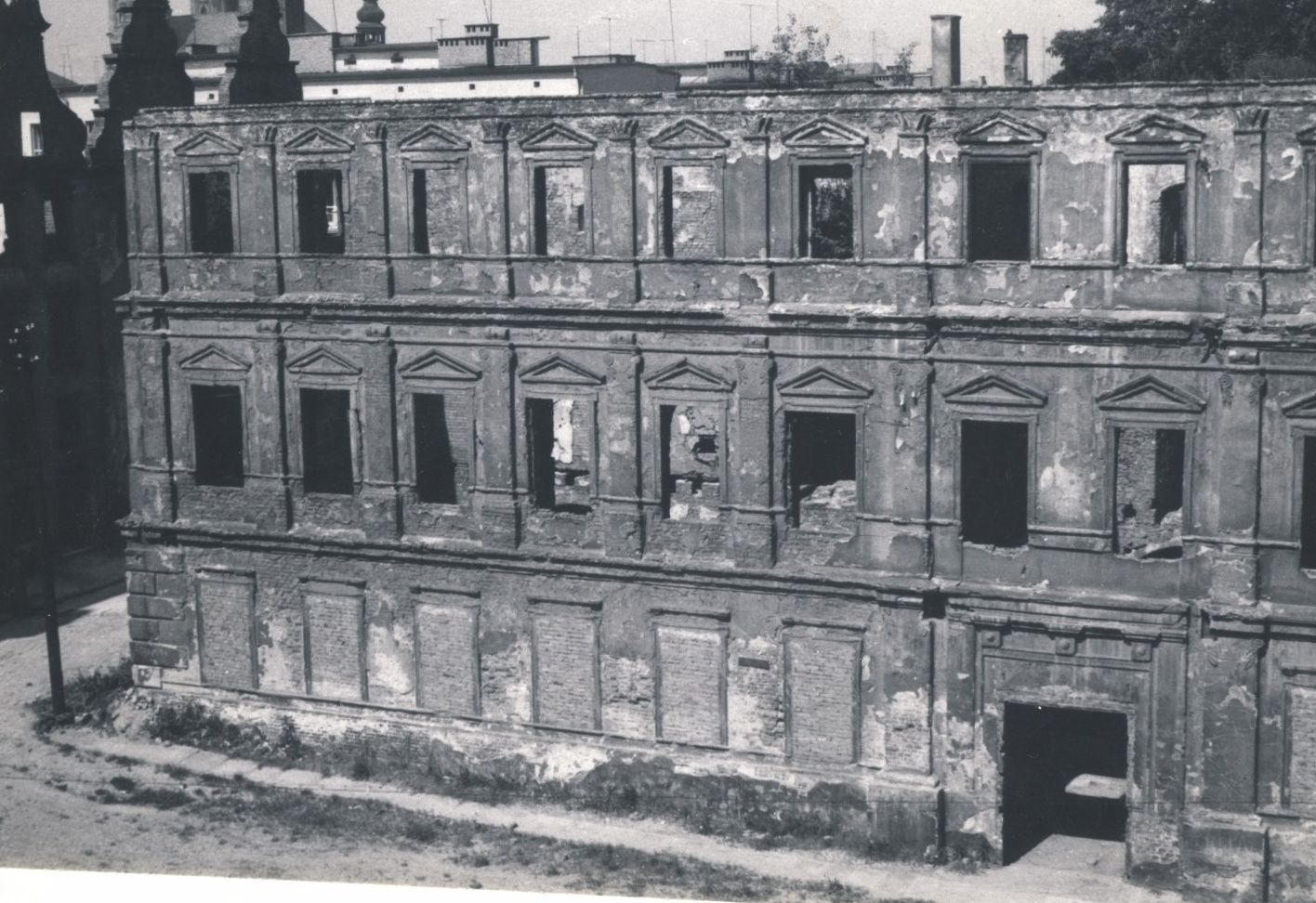 Zdjęcie. Widok przyszłego budynku szkoły przed odbudową ze zniszczeń wojennych. Ruiny pozbawione dachu, stolarki okiennej i drzwiowej. Mury wypalone. Dolne okna są zamurowane. W środku przez wnęki okienne widać brak stropów pomiędzy kondygnacjami.