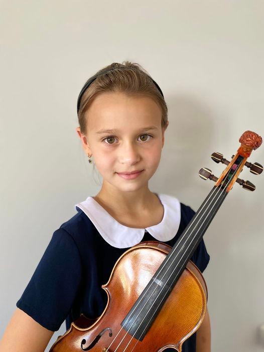 Na zdjęciu znajduje się uśmiechnięta dziewczynka w granatowej sukience z kołnierzykiem. W rękach trzyma skrzypce. 