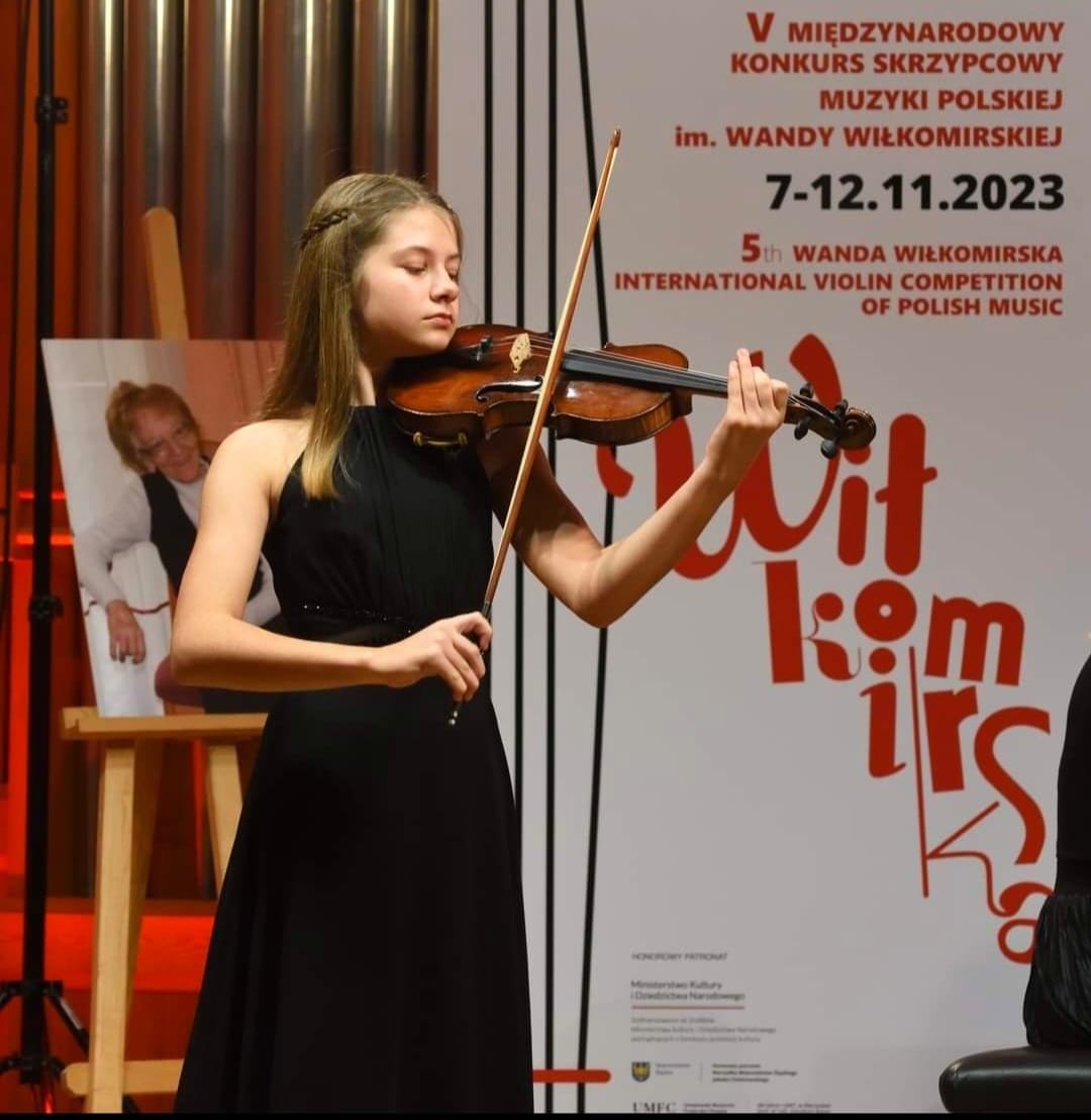 Na zdjęciu widoczna uczennica Agata Kostanowicz grająca na skrzypcach, ubrana w długą czarną sukienkę. W tle widoczny baner konkursowy oraz zdjęcie patronki konkursu - Wandy Wiłkomirskiej. 