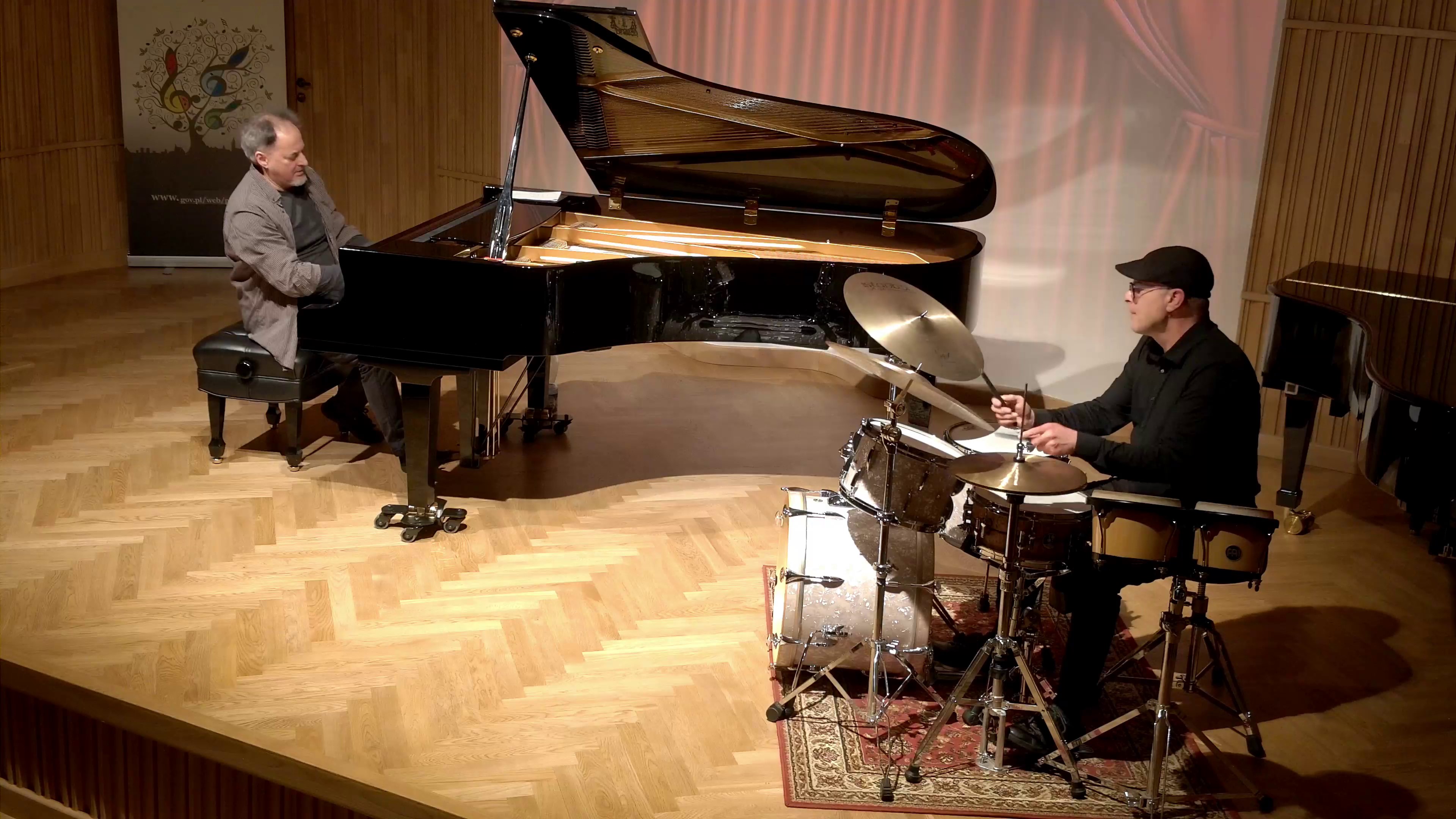 Zdjęcie. Na estradzie sali koncertowej występują: pianista jazzowy grający na dużym czarnym fortepianie oraz perkusista na zestawie perkusyjnym.