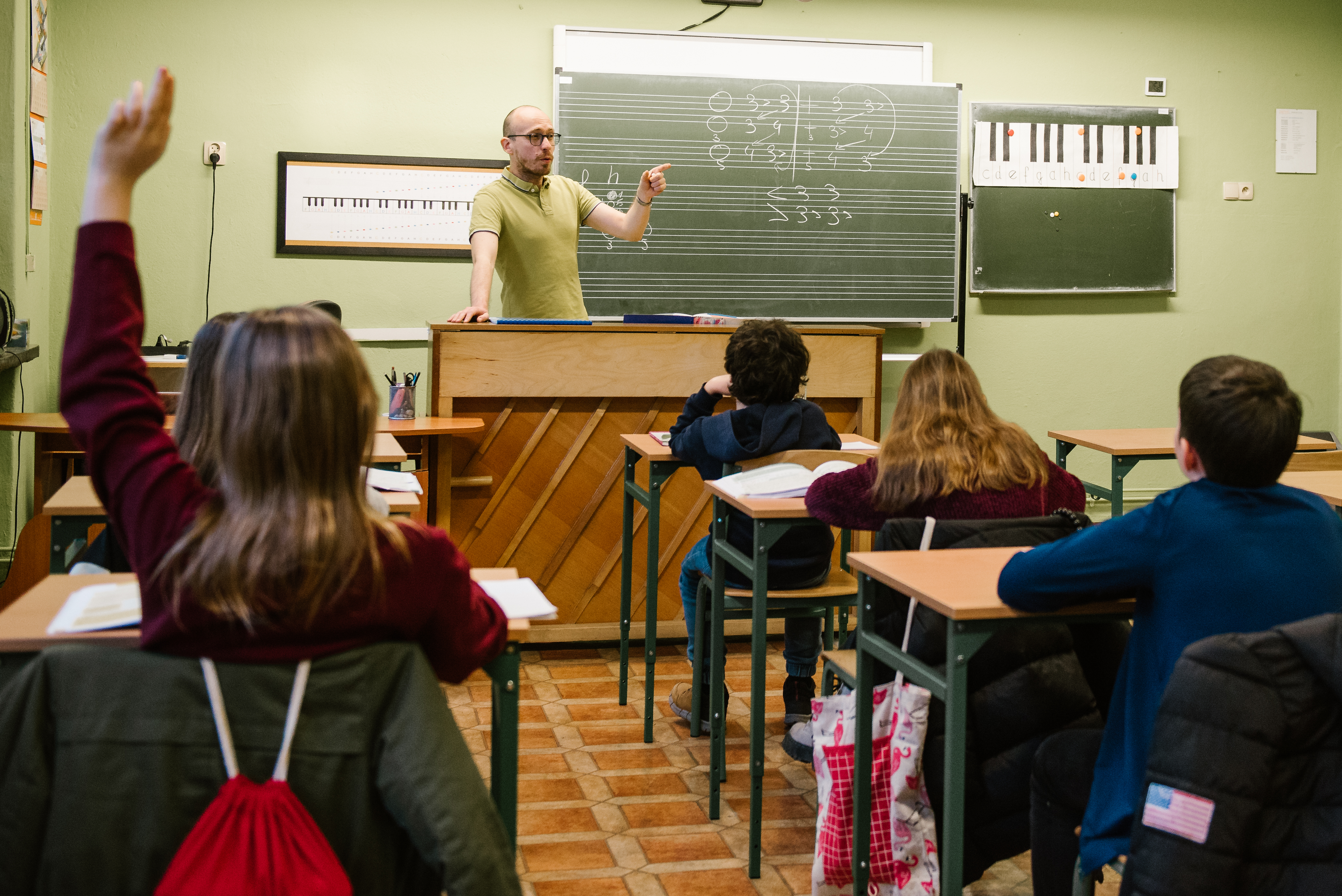 Na zdjęcie znajduje się nauczyciel Dariusz Latosi oraz grupa uczniów. Nauczyciel stoi oparty przy pianinie z uniesioną lewą ręką, uczniowie siedzą w ławkach , na których znajdują się książki i zeszyty. Na ścianie w tle wiszą tablice oraz meloplast. 