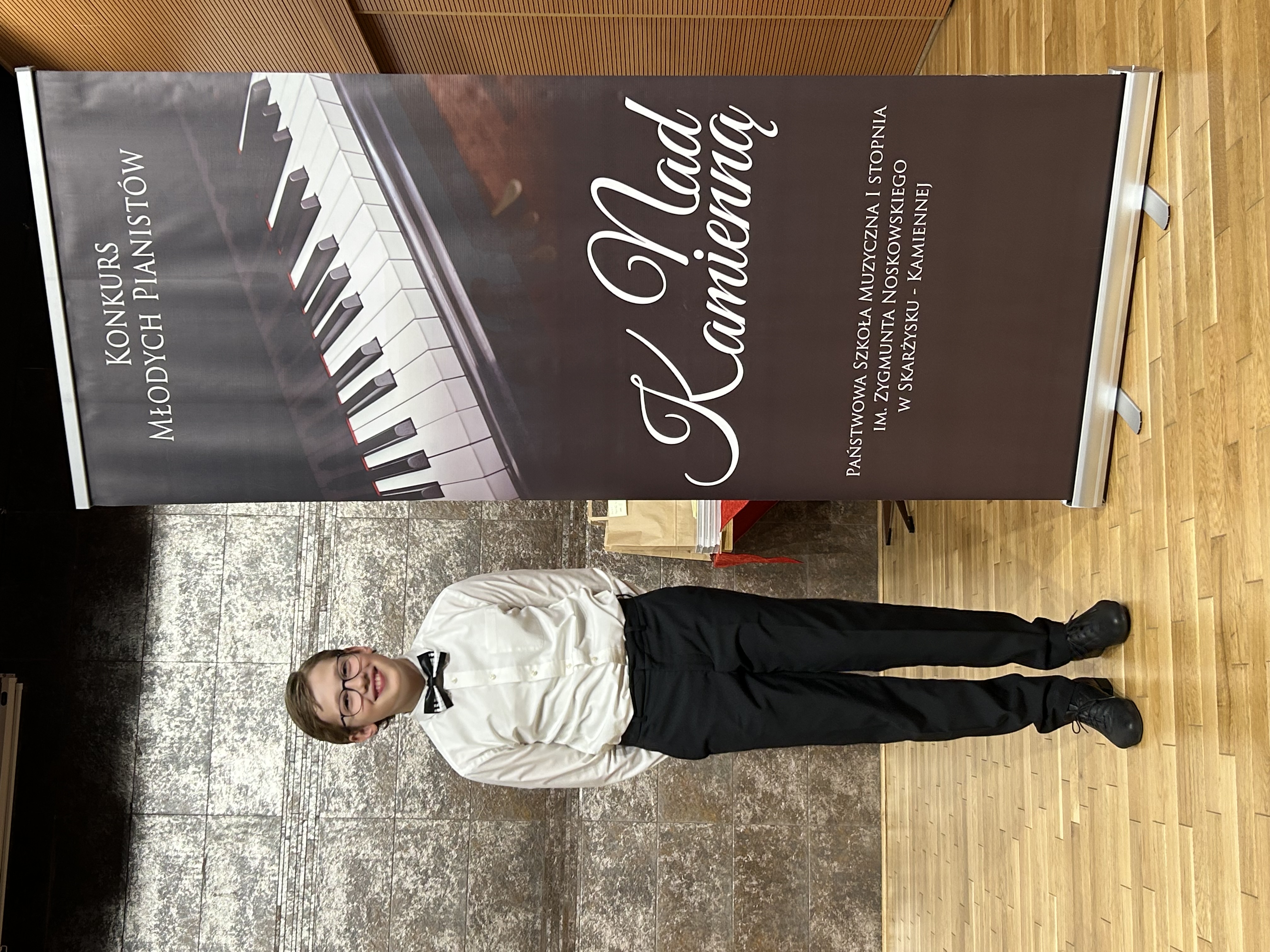 Na scenie Gabriel Kuizinas ubrany na galowo stoi przy bannerze reklamowym konkursu. 