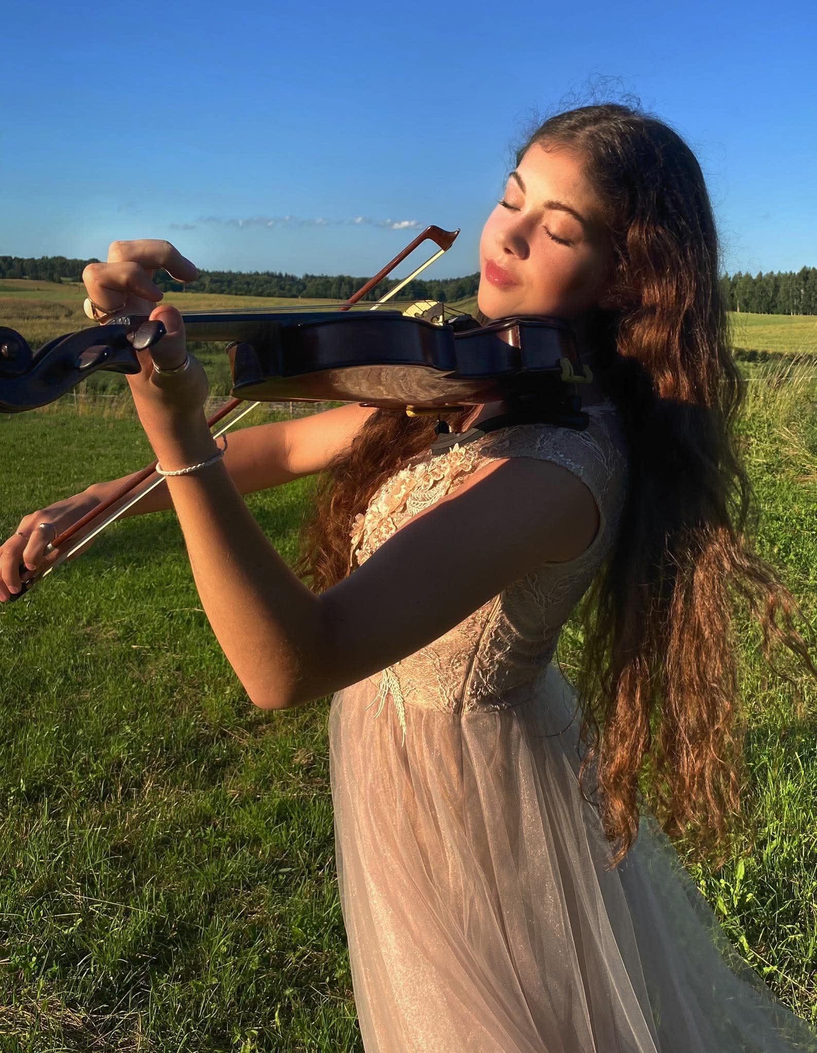 Zdjęcie dziewczyny ze skrzypcami na łące. Młoda dziewczyna, z rozpuszczonymi włosami i zwiewnej sukience, gra z zamkniętymi oczami.