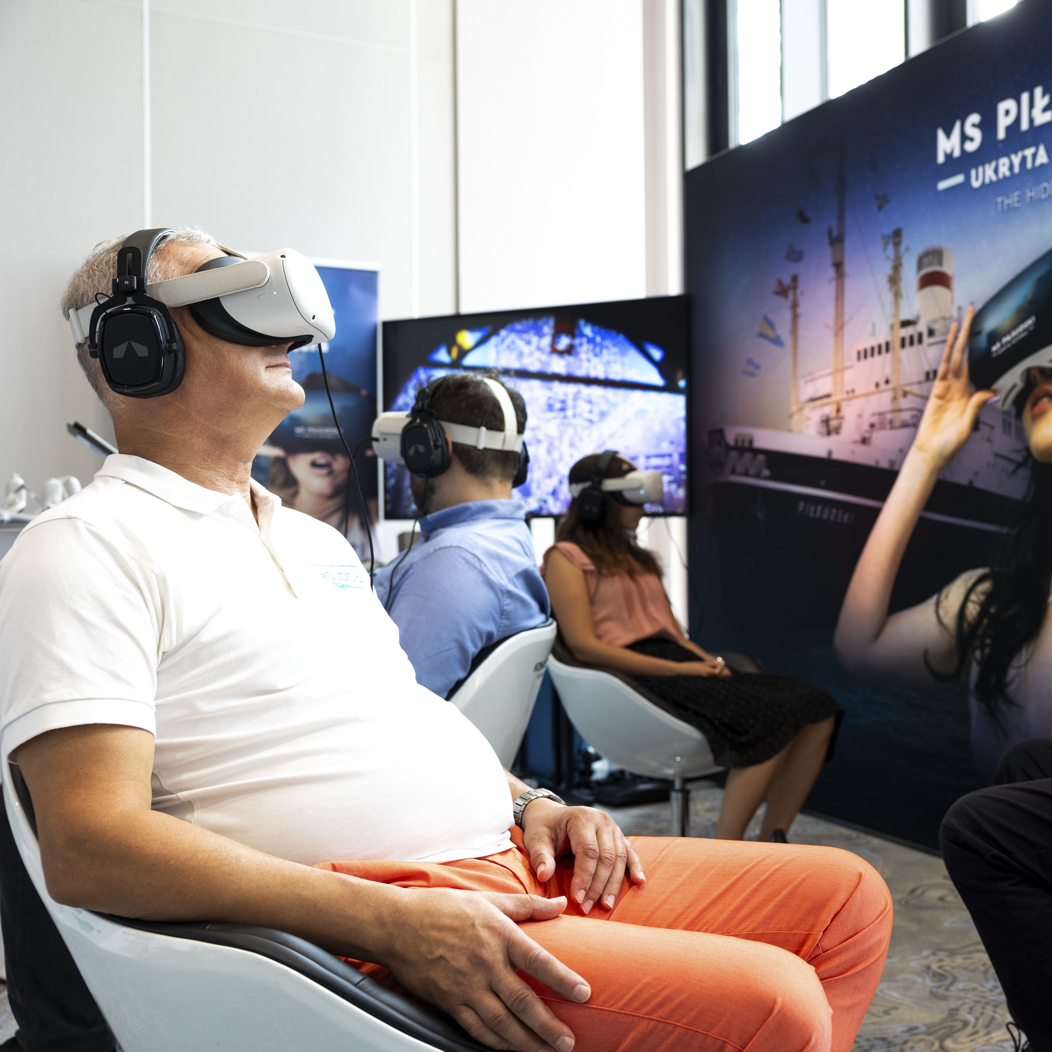 Trzy osoby siedzą na fotelach z założonymi okularami i słuchawkami do Virtual Reality