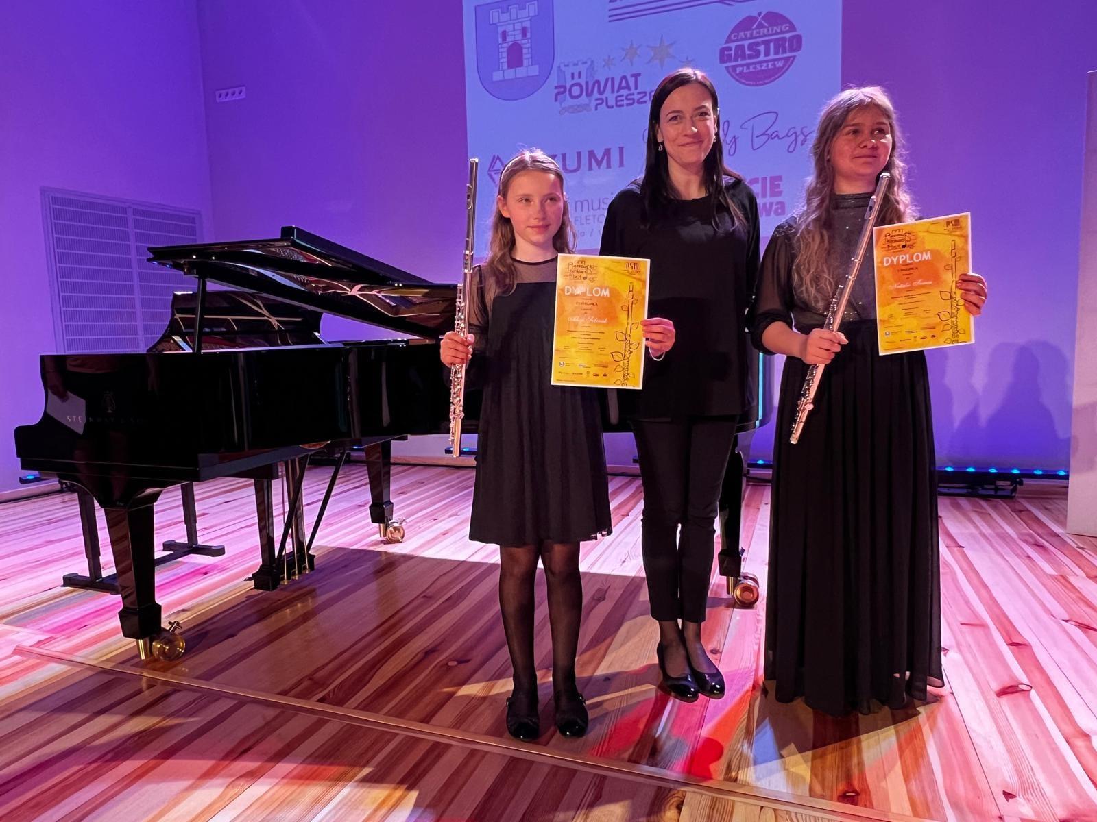 Na zdjęciu znajdują się dwie uczennice w czarnych sukienkach, w rękach trzymają dyplomy konkursowe oraz flety. Między nimi znajduje się Pani akompaniatorka. W tle znajduje się czarny fortepian.