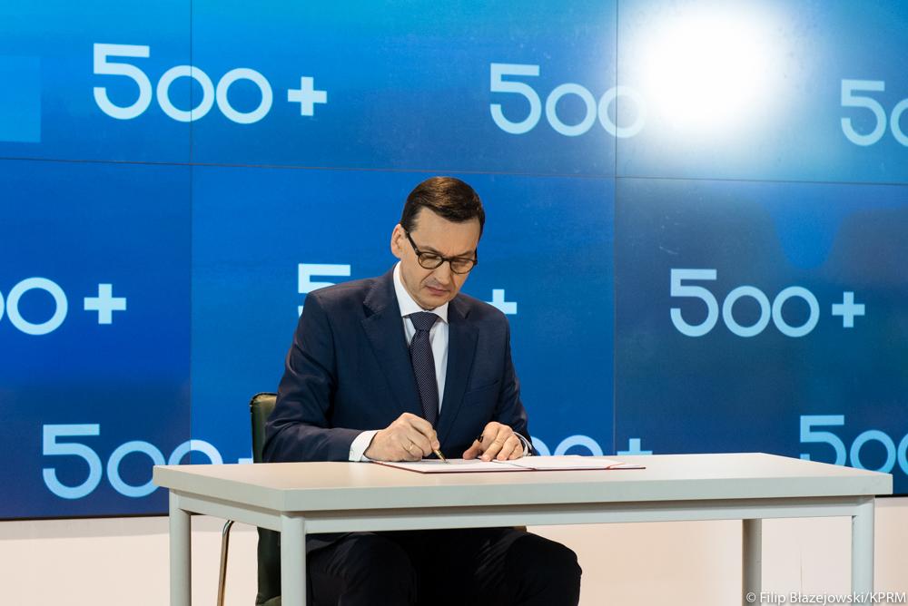 Premier Mateusz Morawiecki podpisuje list na tle loga 500+.