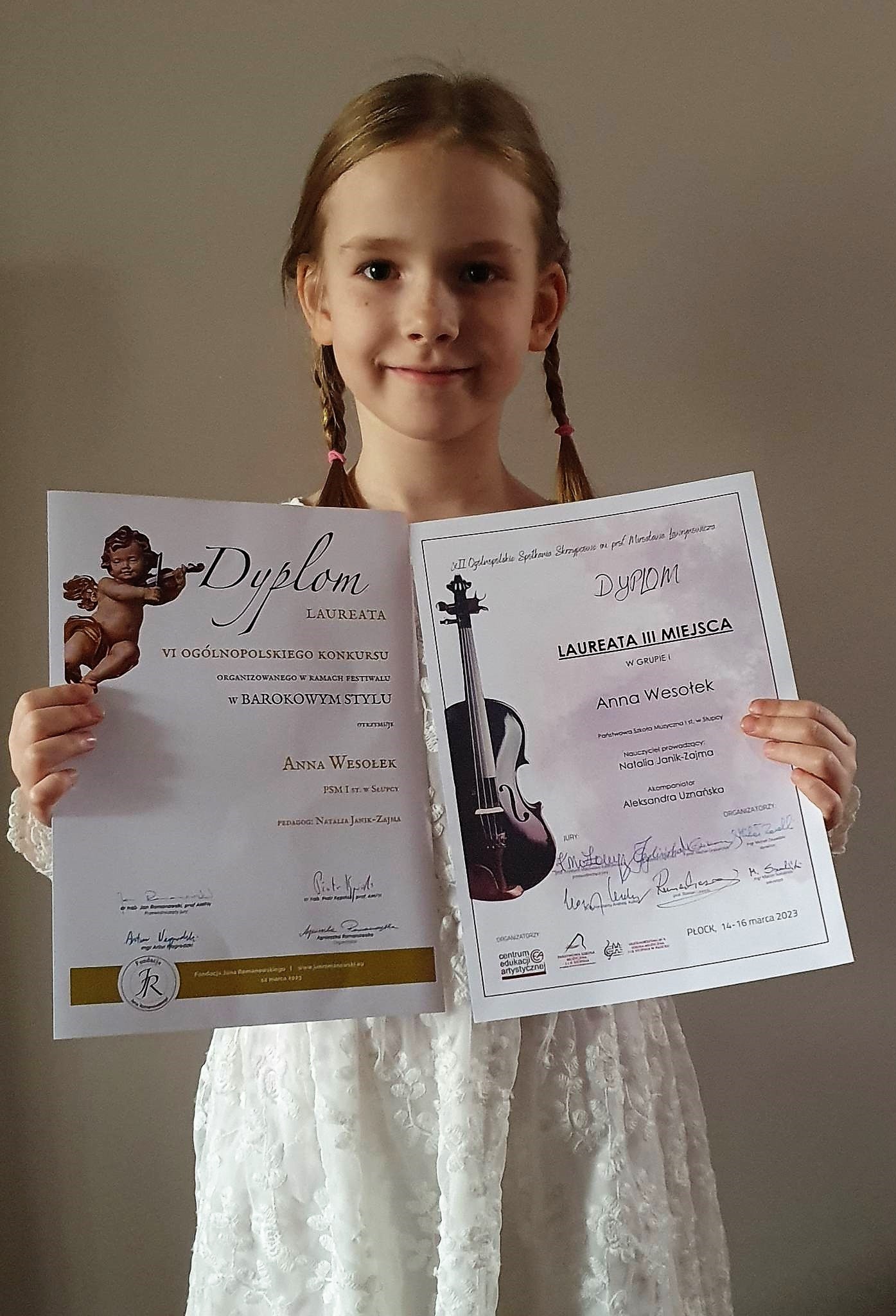Dziewczynka w białej sukience trzymająca 2 dyplomy konkursowe, jasnobrązowe tło