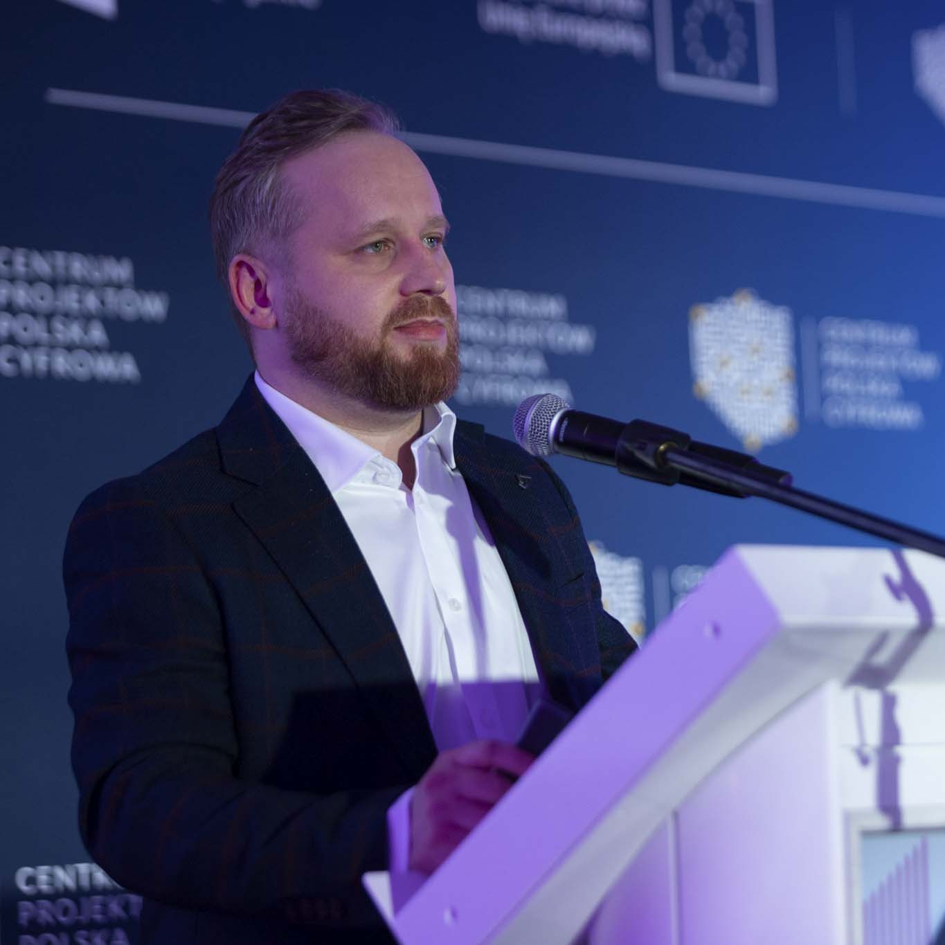 Przy mównicy stoi Pan Kamil Gaweł. Wzrok kieruje w stronę uczestników konferencji. Mówi do mikrofonu.