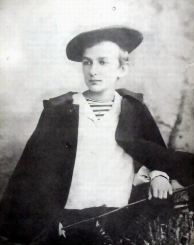 Czarno-biały portret Szymanowskiego w wieku 15 lat. Na zdjęciu młodzieniec w ciemnym płaszczu, jasnej koszuli i wystającym spod niej podkoszulku w marynarskie paski. Na głowie duży, ciemny beret.