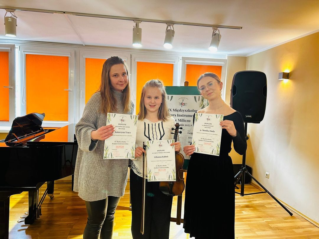 Na zdjęciu widoczna jest uśmiechnięta uczennica stojąca pomiędzy dwiema nauczycielkami naszej szkoły. Wszystkie trzymają w rękach dyplomy. Zdjęcie zostało zrobione w sali koncertowej Szkoły Muzycznej w Miliczu.