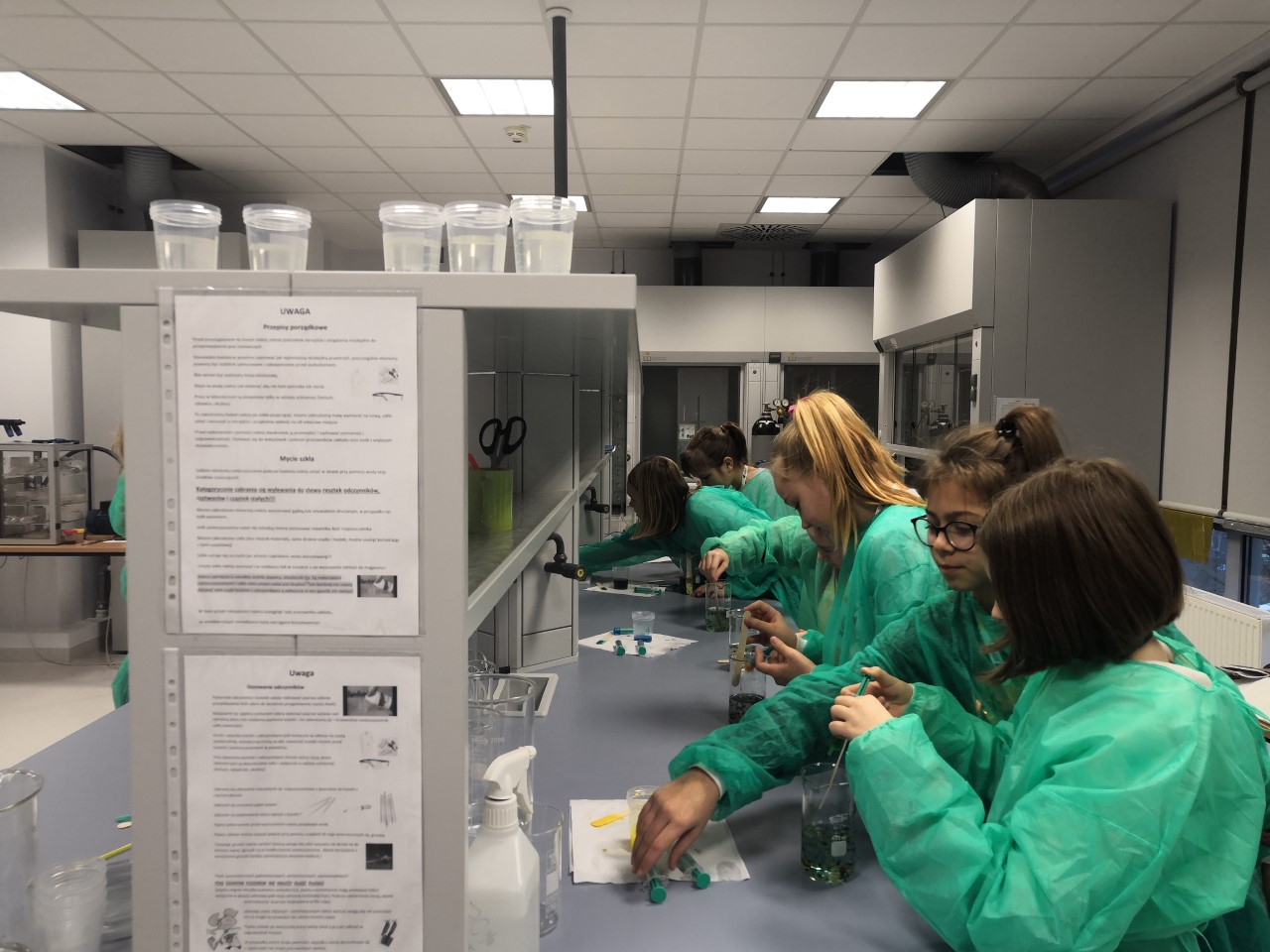 Zajęcia w laboratorium. Siedem nastolatek w zielonych fartuchach, stojąc w rzędzie przy długim blacie, wlewa do menzurek płyn za pomocą strzykawek