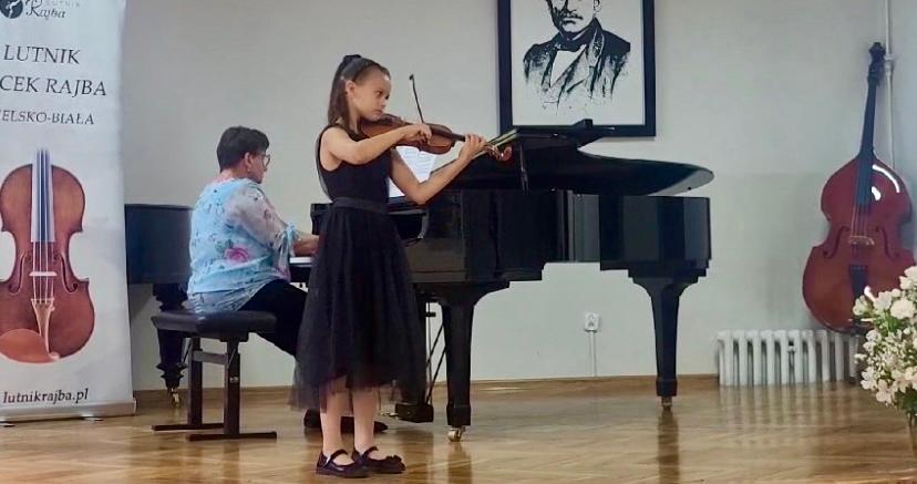 Na zdjęciu znajduje się dziewczynka w czarnej sukience grająca na skrzypcach. Za nią znajduje się akompaniatorka przy fortepianie. W tle widoczny baner konkursowy, kontrabas oraz obraz. 