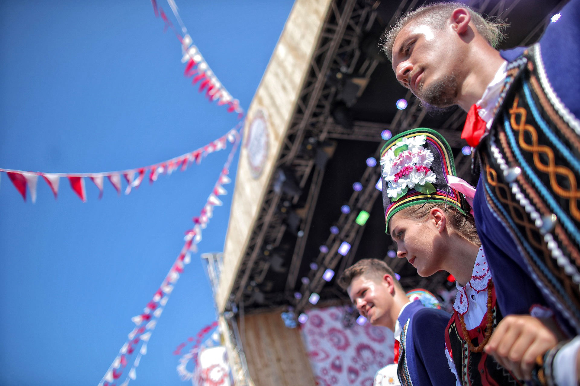 Dwóch mężczyzn i kobieta ubrani w stroje ludowe, w tle scena