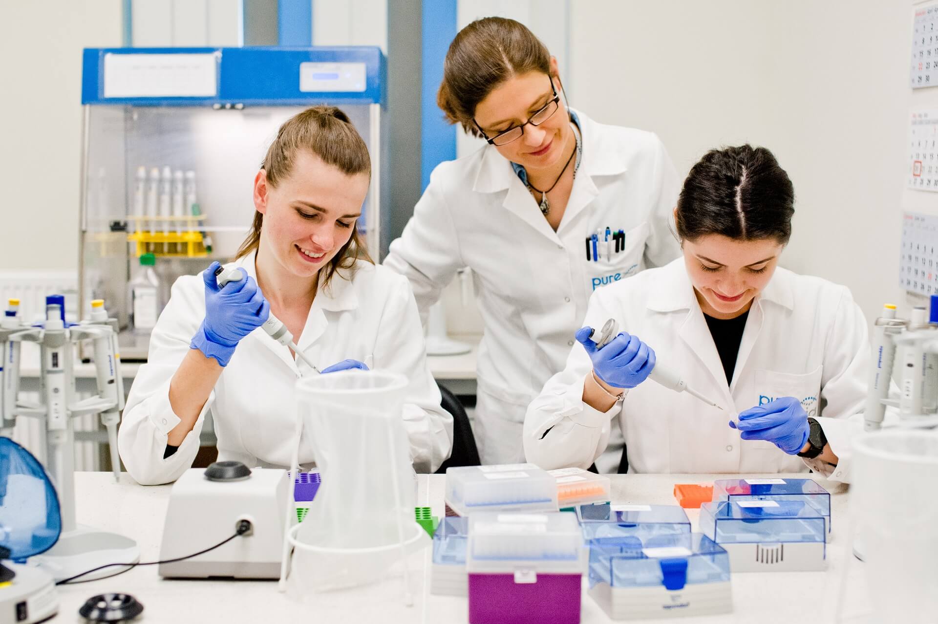 Trzy uśmiechnięte kobiety w białych kitlach, pracujące w laboratorium