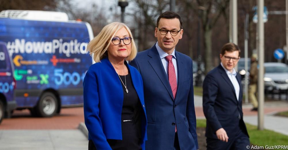 Premier Mateusz Morawiecki i minister Joanna Kopcińska idą na tle busów Nowej Piątki.