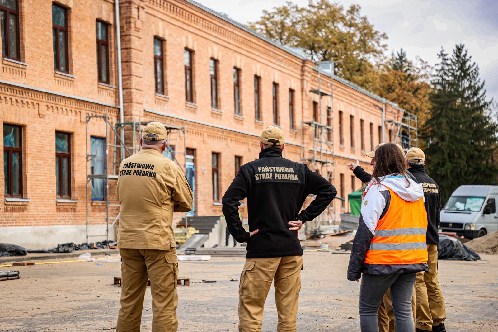 4 strażaków i 1 kobieta w cywilnym ubraniu stoją przed remontowanym budynkiem z czerwonej cegły