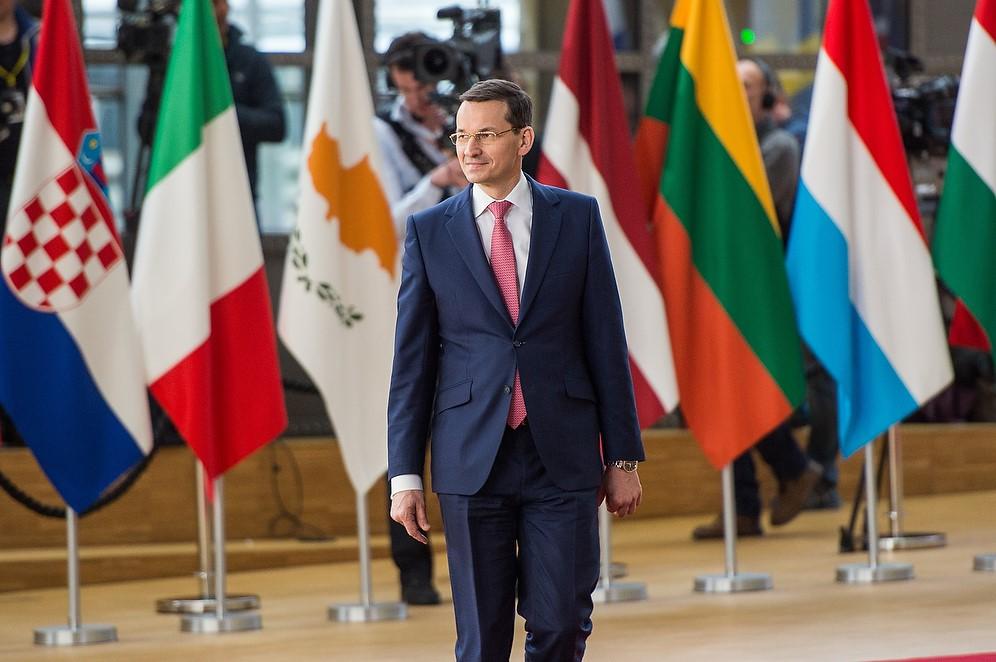 Premier Mateusz Morawiecki idzie na tle flag.