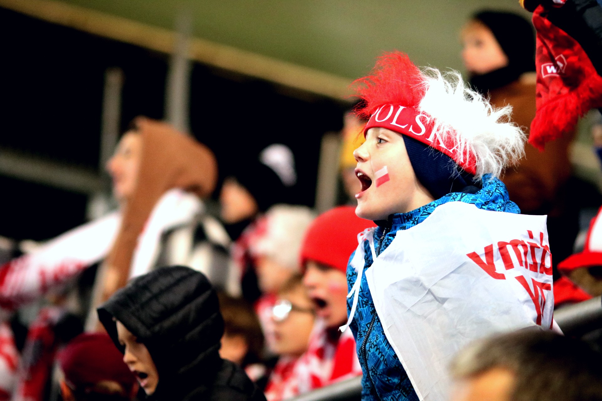 Dziecko w niebieskiej kurtce stoi i krzyczy, ma biało-czerwone paski na policzkach i biało-czerwoną puchatą czapkę z napisem Polska.