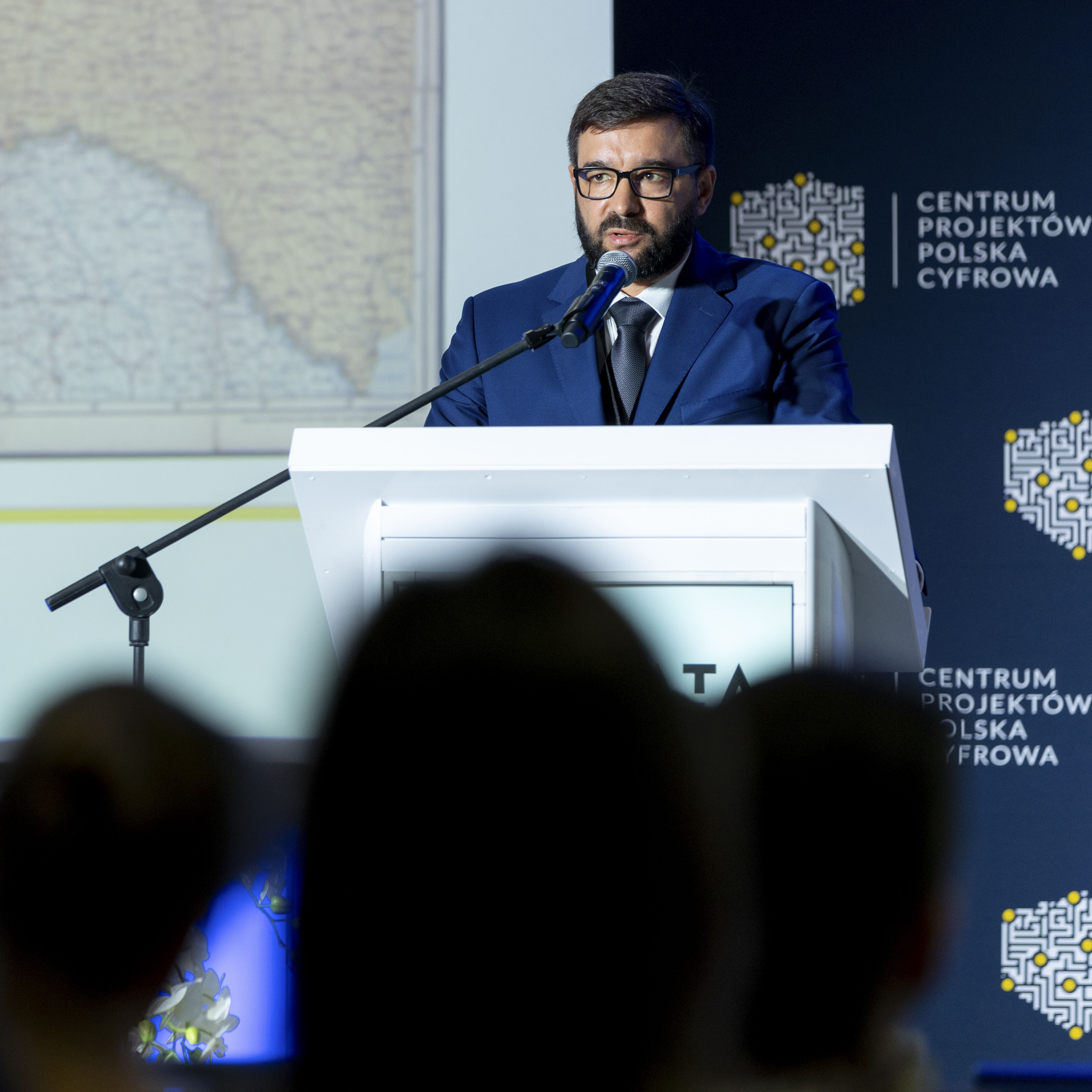 Przy mównicy stoi Pani Agnieszka Krauzowicz. W ręku trzyma mikrofon patrząc na siedzących przed nią uczestników konferencji.