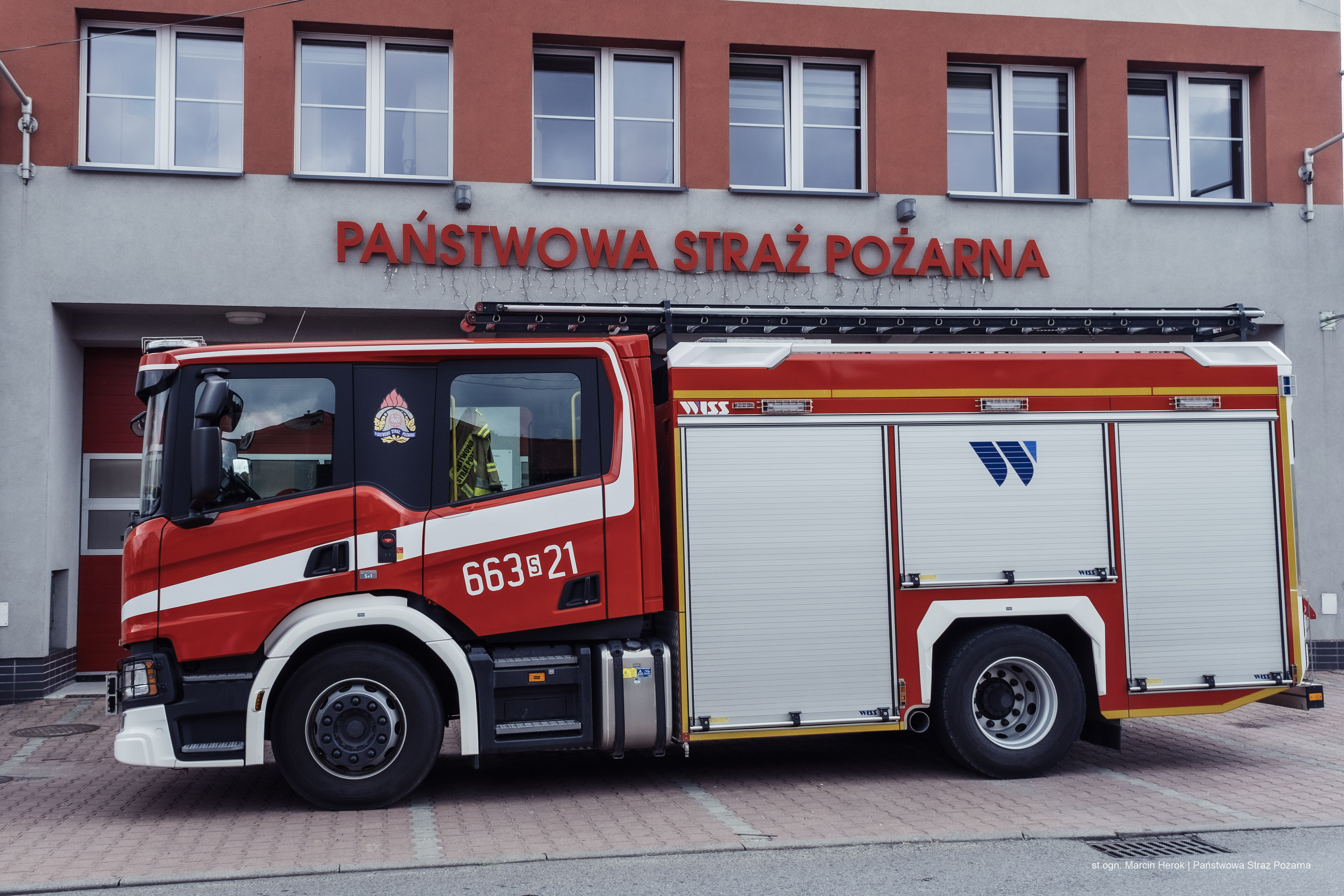 Zdjęcie przedstawia średni samochód ratowniczo-gaśniczy. W tle budynek Jednostki Ratowniczo-Gaśniczej w Rydułtowach.