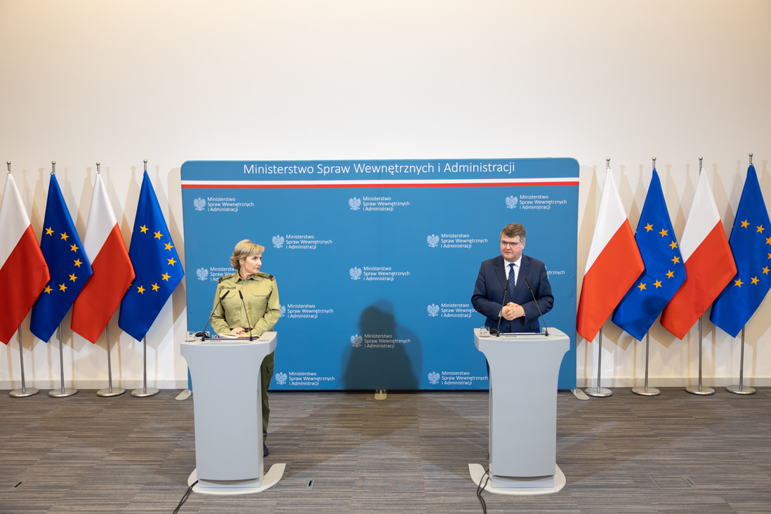 Na zdjęciu widać wiceministra Macieja Wąsika i gen. bryg. SG Wioletę Gorzkowską stojących za mównicami w trakcie konferencji prasowej. W tle widać ściankę MSWiA, flagi Polski i UE.
