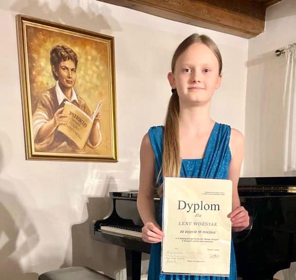 Na zdjęciu widoczna jest uśmiechnięta uczennica ubrana w niebieską sukienkę, trzymająca dyplom z konkursu. W tle widoczny jest wiszący złoty obraz oraz czarny fortepian.