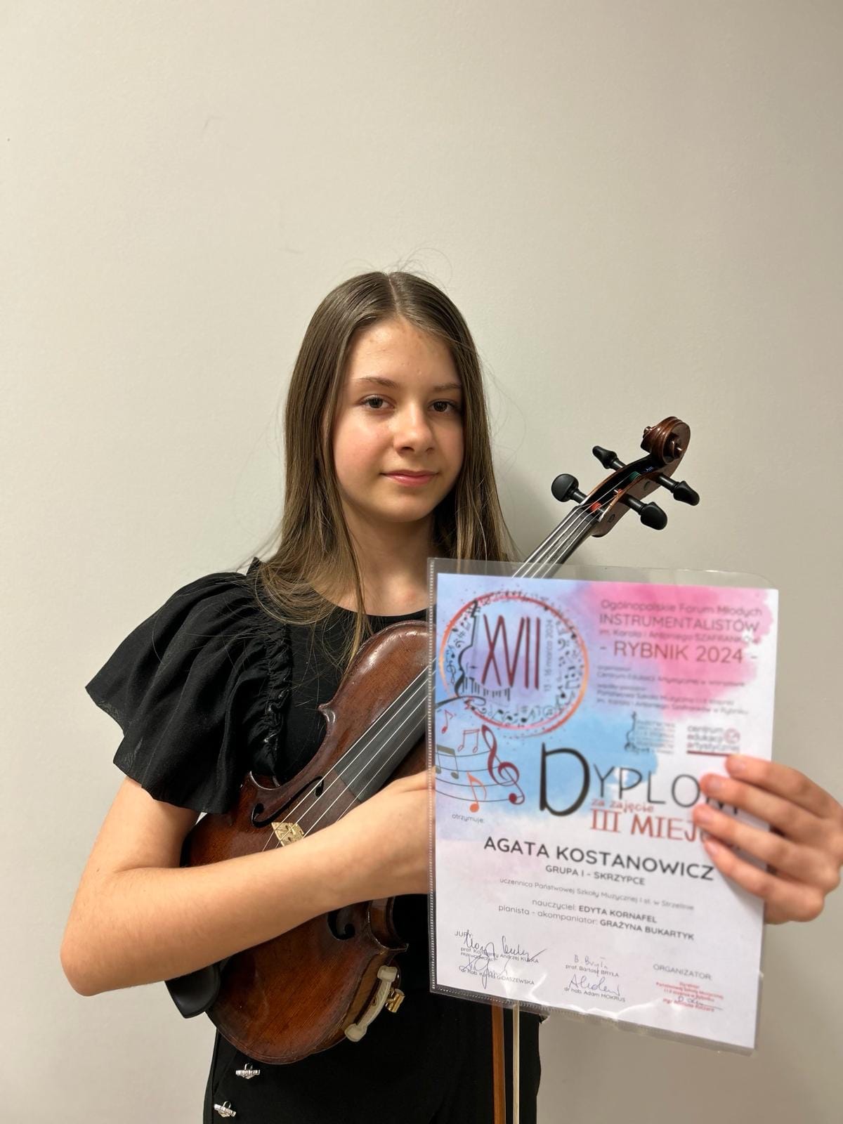 Na zdjęciu znajduje się uczennica w czarnej sukience. W prawej ręce trzyma skrzypce, w lewej dyplom konkursowy. 