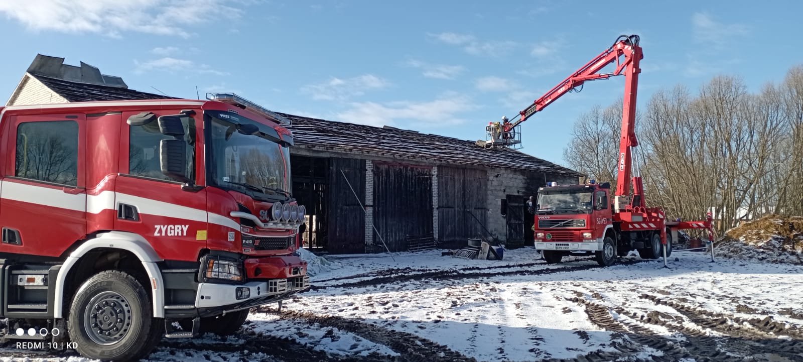 Po prawej stronie zdjęcia znajduje się podnośnik pożarniczy, który bierze udział podczas usuwania skutków nawałnicy. Po środku znajduje się budynek inwentarsko-składowy z zerwanym poszyciem dachu. Po lewej stronie znajduje się pojazd pożarniczy OSP KSRG. 