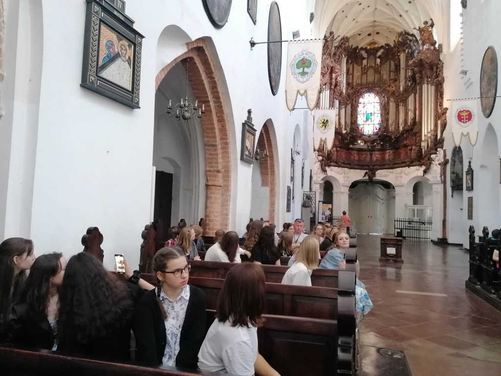 Kolorowe zdjęcie. Wnętrze kościoła - ławki, w nich siedzący ludzie. W tle barokowe organy.