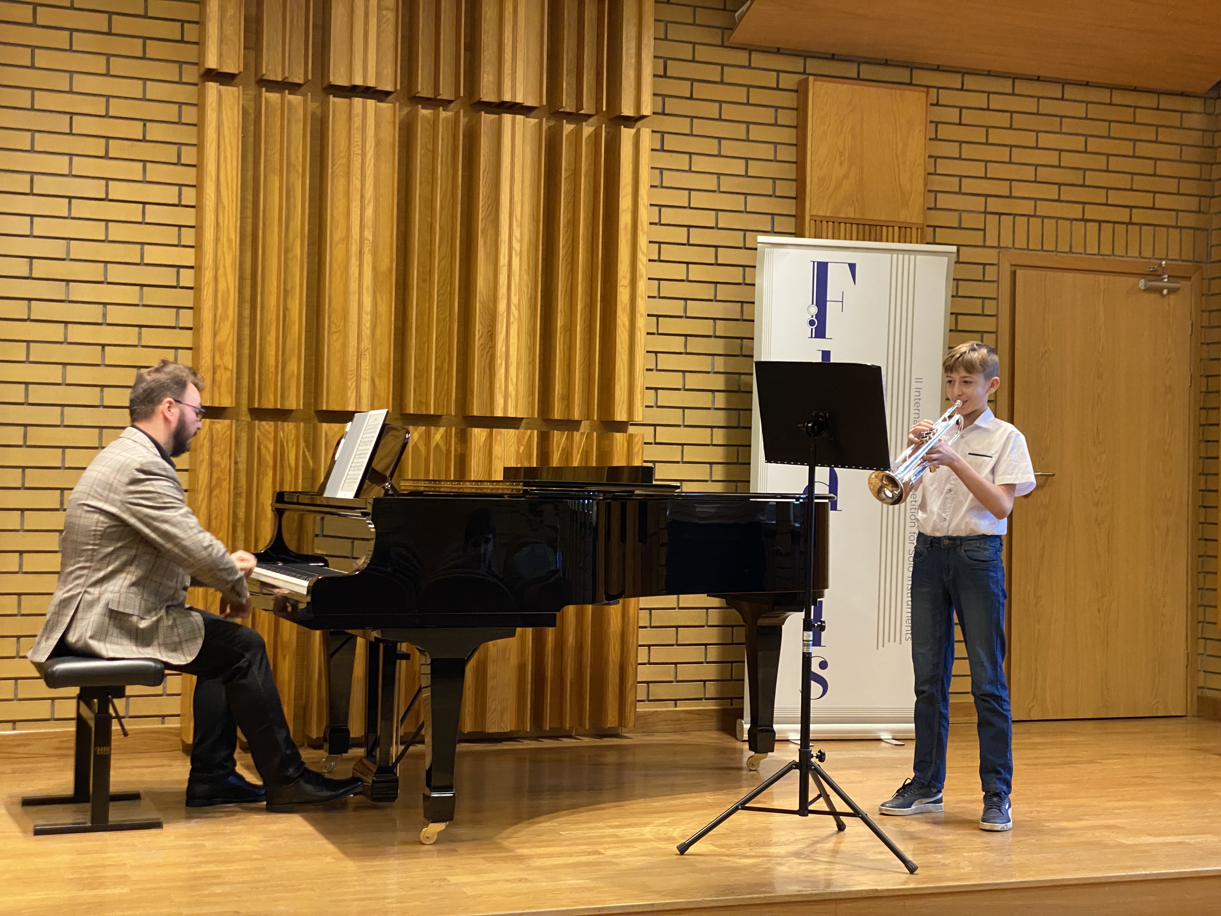 Zdjęcie przedstawia scenę w brązowym kolorze, na której stoi chłopiec ubrany na galowo i gra na srebrnej trąbce. Przed nim stoi czarny pulpit a obok czarny fortepian, na którym gra mężczyzna ubrany w czarne spodnie i brązową marynarkę w kratkę. 