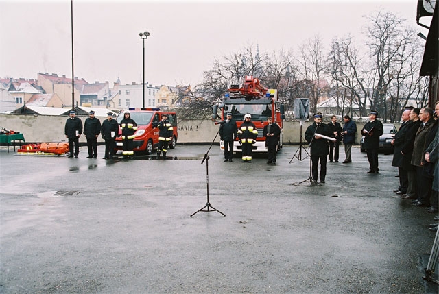 Na placu JRG nr 1 Nowy Sącz stoją przekazywane samochody. Obok samochodów stoją strażacy. Komendant Miejski przemawia do zgromadzonych gości.