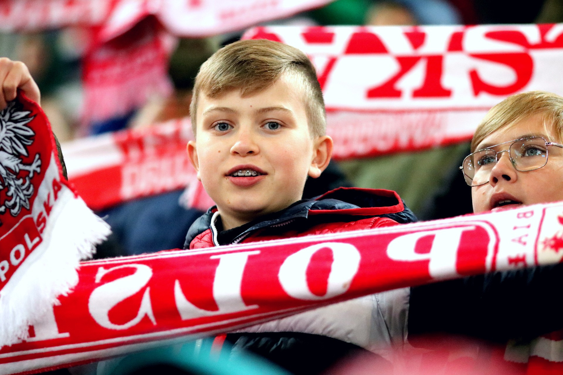 Chłopiec na trybunie stadionu trzyma rozpostarty biało-czerwony szalik z napisem Polska, za nim widać inne takie szaliki.