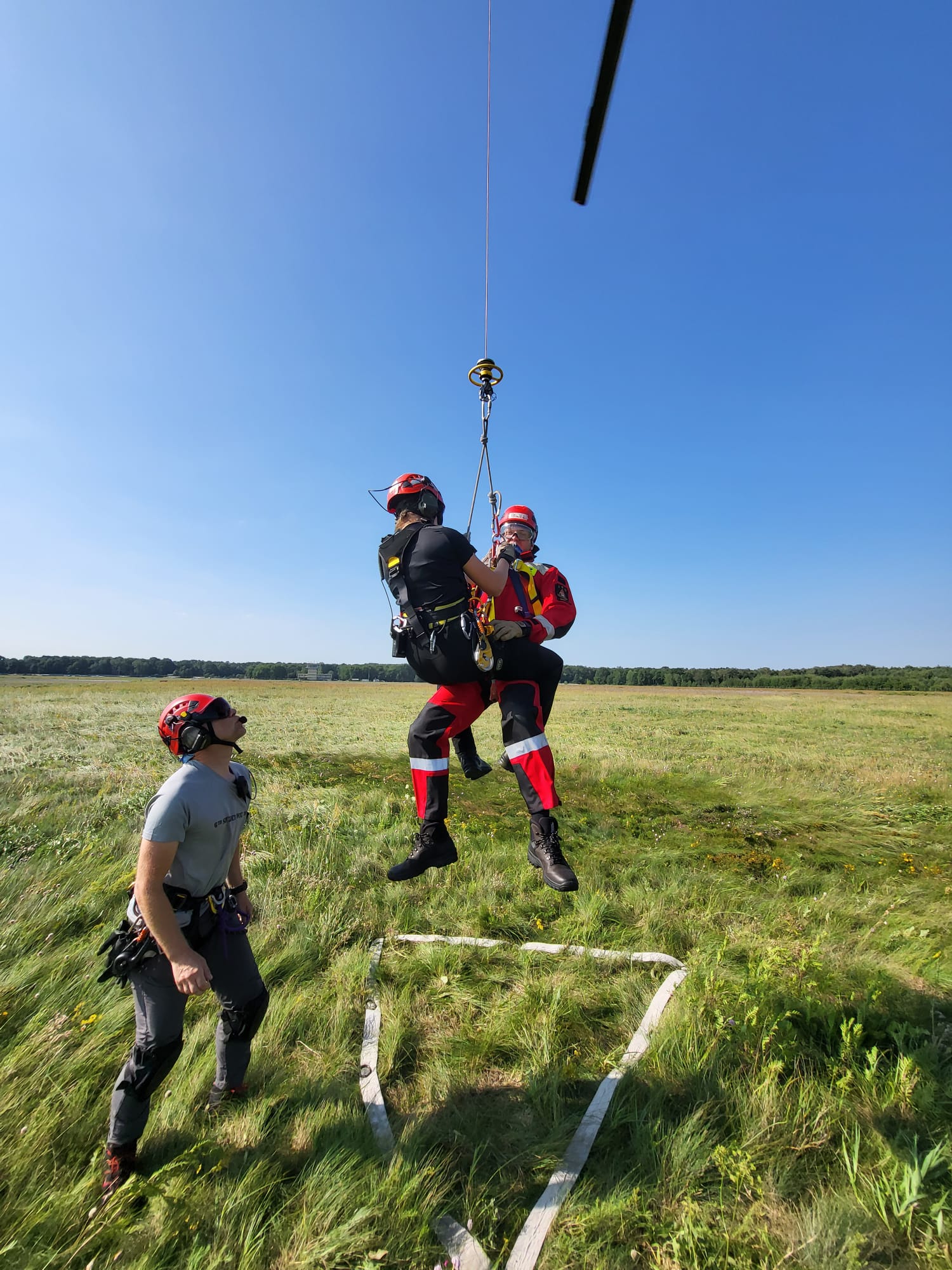 Dwóch ratowników zamocowanych na linie uniesieni lekko nad ziemią, obok stoi 3 ratownik patrząc w górę (na śmigłowiec którego na zdjęciu nie widać) - ćwiczenia