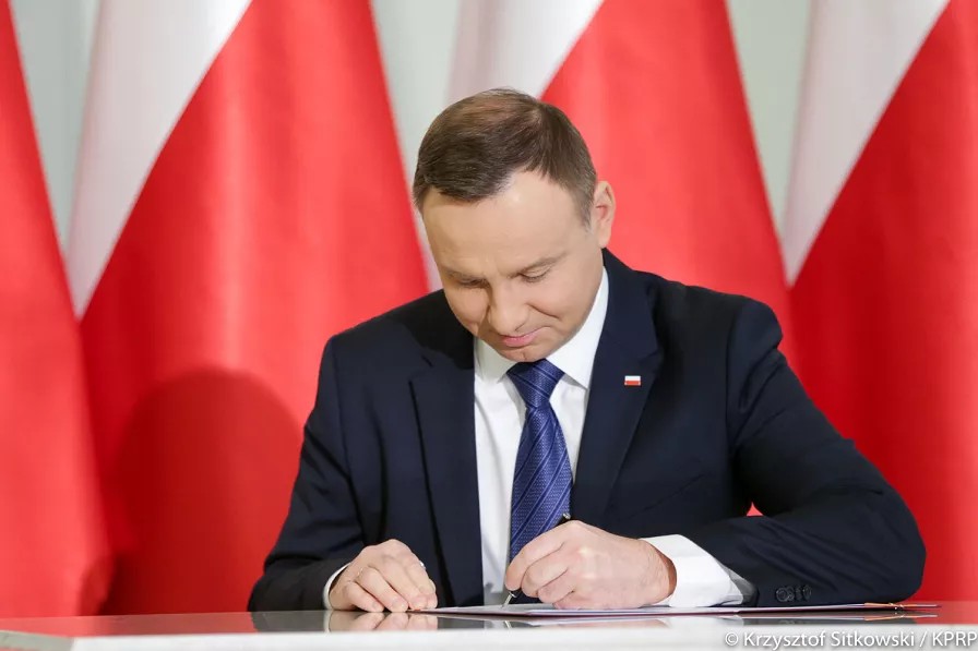 Prezydent Andrzej Duda, który podpisuje dokument. W tle polskie flagi.