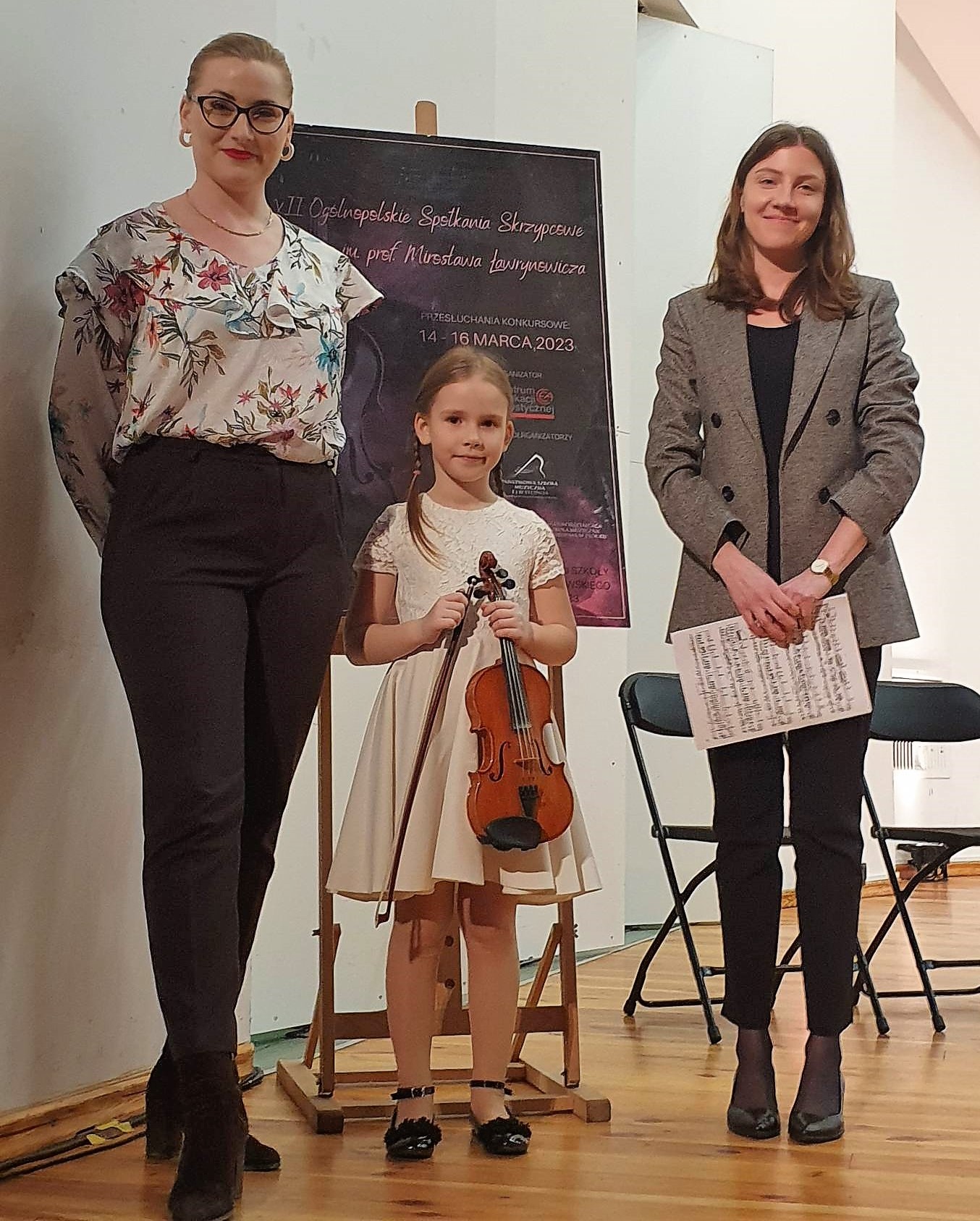 Trzy osoby na scenie, w środku dziewczynka ze skrzypcami, za nimi plakat konkursowy, wszystko na tle białej ściany