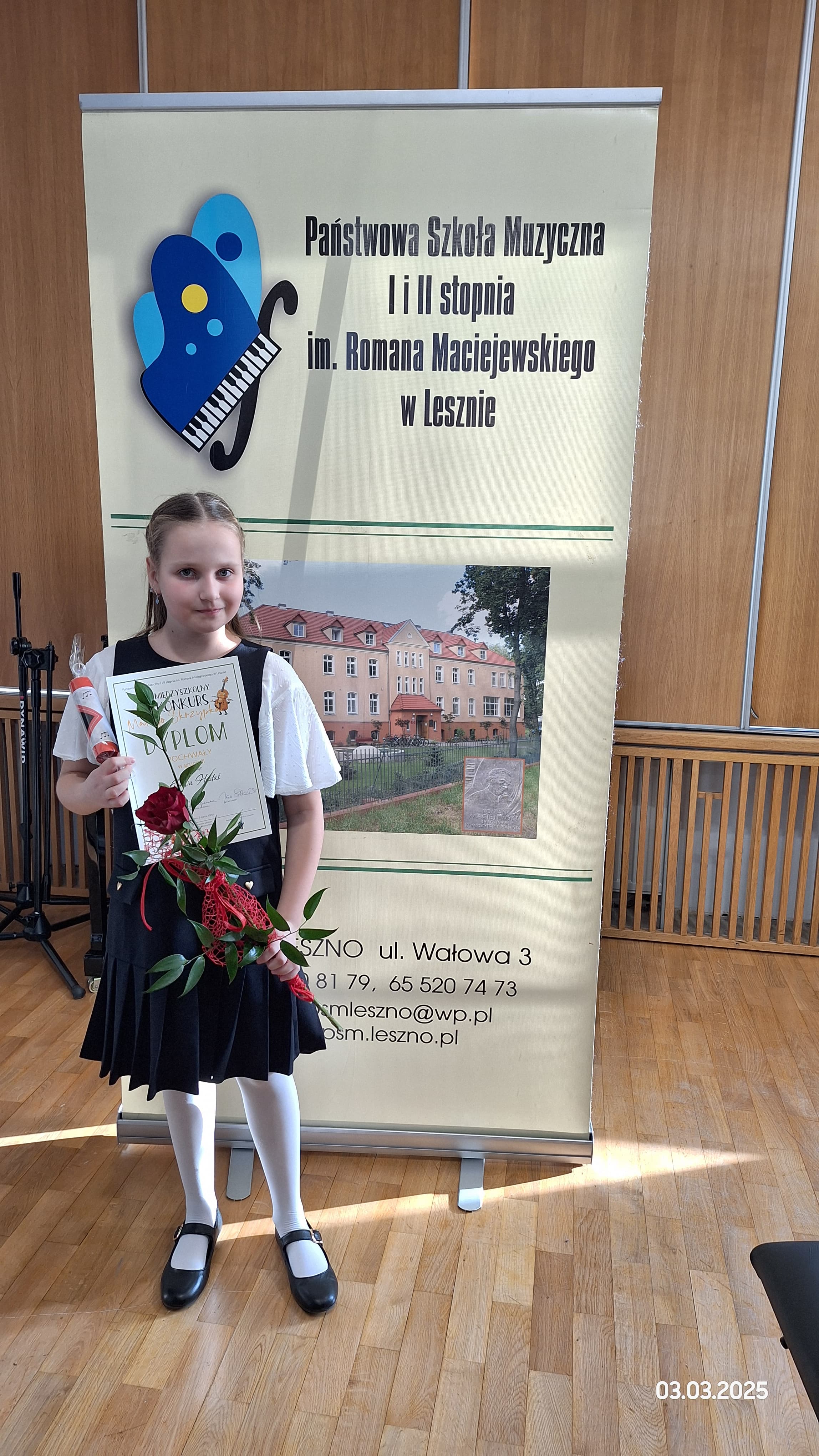 Uczennica jest elegancko ubrana. Stoi przed banerem reklamującym Państwową Szkołę Muzyczną w Lesznie. W rękach trzyma różę, dyplom i nagrodę. 
