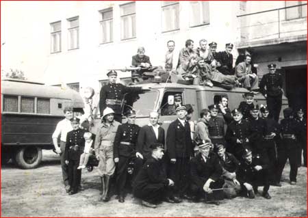 Zdjęcie historyczne KP PSP w Kozienicach przedstawiające wozy strażackie przed budynkiem komendy, a wokół jednego z nich zgromadzeni są strażacy. Cześć z nich stoi przed wozem, część się o niego opiera, a część siedzi na jego dachu.