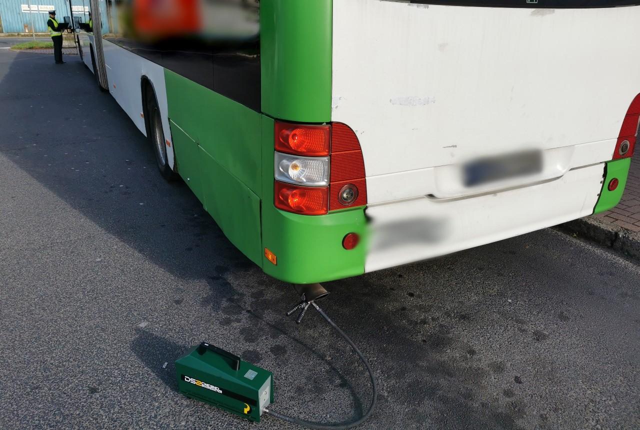Inspektor pobierający dokumenty od kierowcy i dymomierz podłączony do wydechu autobusu.