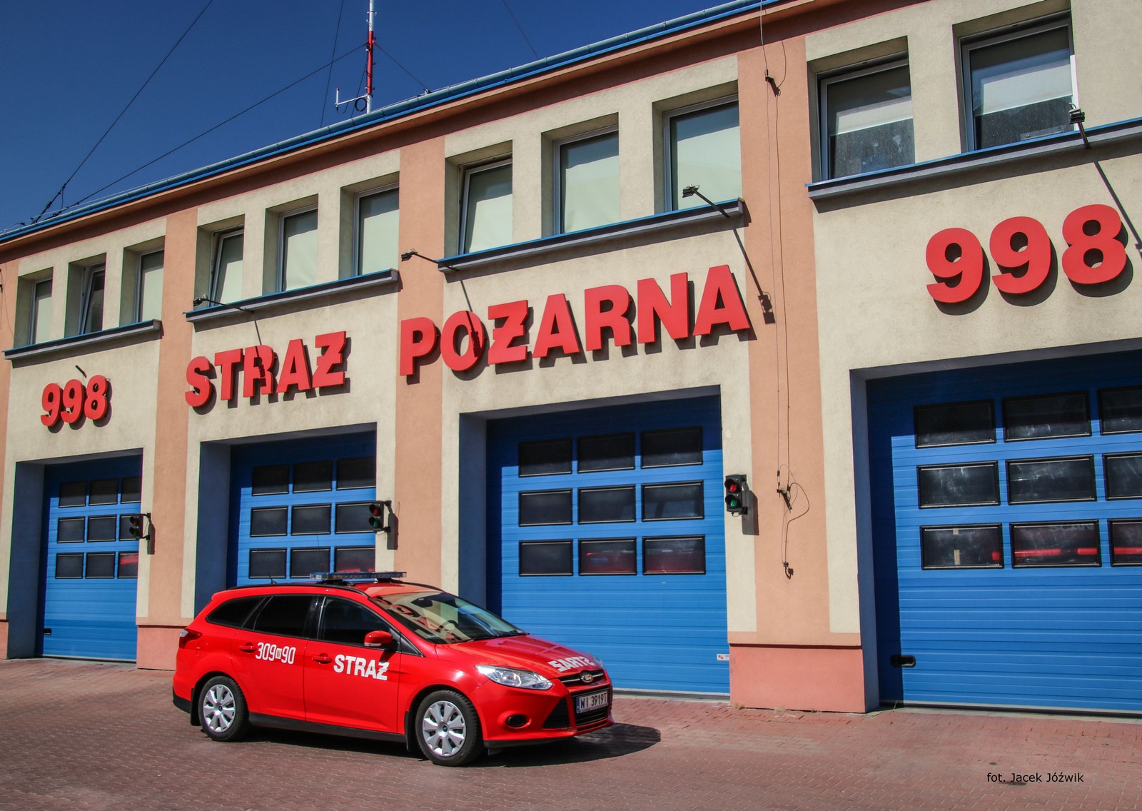 Na pierwszym planie widać czerwony, pożarniczy samochód osobowy. Na boku pojazdu widoczny jest kryptonim operacyjny pojazdu - numer 309W90 oraz napis STRAŻ. Na dachu znajduje się belka z niebieskimi sygnałami ostrzegawczymi. W tle można zobaczyć żółto-pomarańczowy budynek Jednostki Ratowniczo-Gaśniczej, czerwony napis "998 Straż Pożarna" oraz niebieskie bramy garażowe.