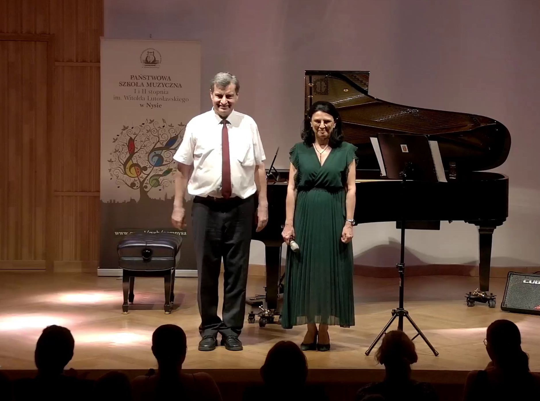 Zdjęcie. na estradzie sali koncertowej przed publicznością stoją Pani Wicedyrektor i pan Dyrektor. Za nimi w tle czarny fortepian i baner PSM Nysa.