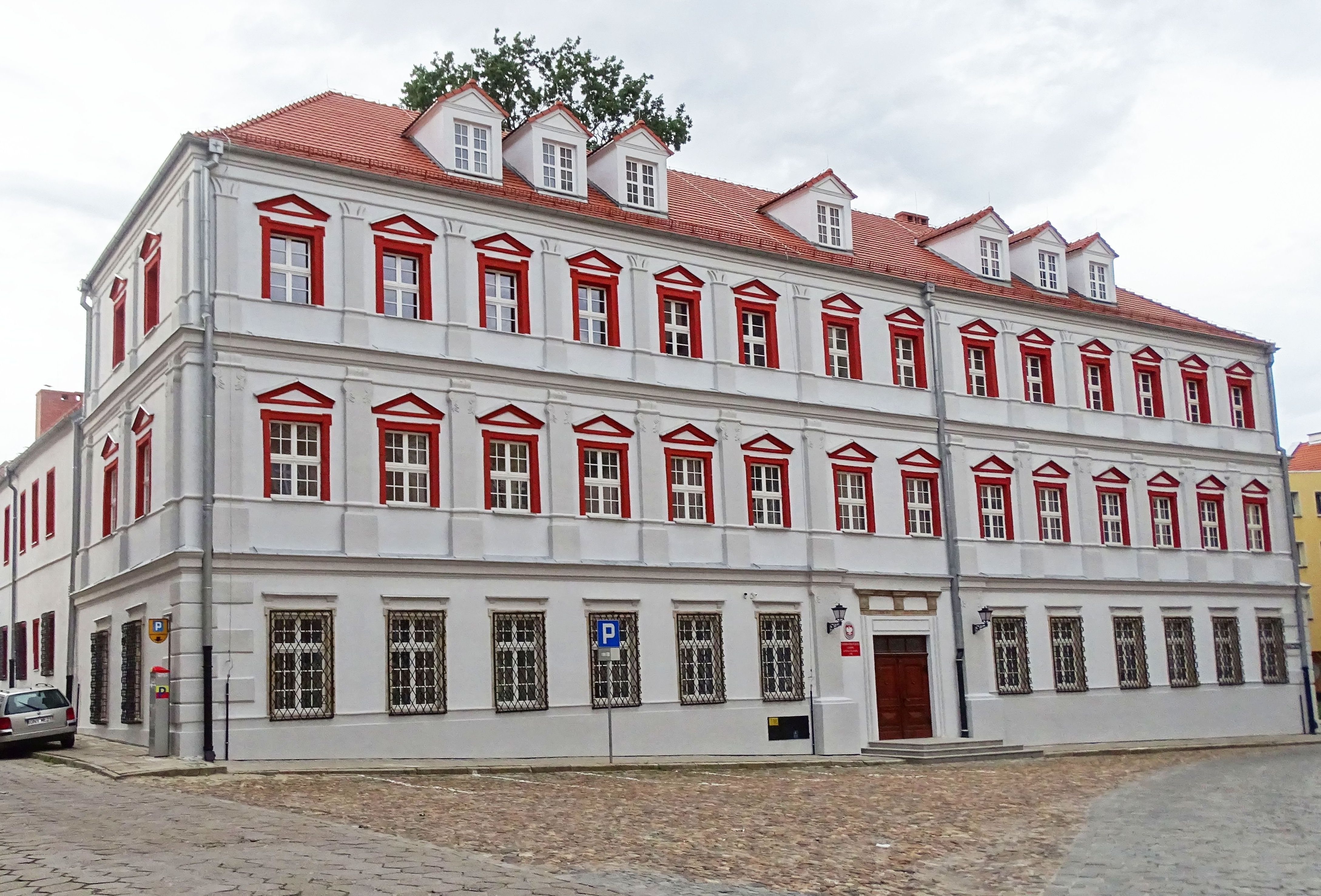 Zdjęcie. Budynek PSM w Nysie - widok po przeprowadzonej renowacji elewacji. Kolor elewacji szary, obramowania okien piętra pierwszego i drugiego czerwone. Okna dolne są chronione czarnymi kratami. Na dachu widać siedem lukarn. Przed budynkiem widać fragment zabytkowej wybrukowanej powierzchni Rynku Solnego. 