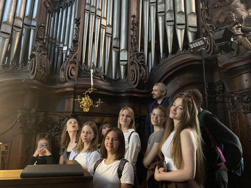 Zdjęcie kolorowe. Grupa uczniów przy stole gry organów kościelnych. W tle płaskorzeźby i piszczałki organowe.