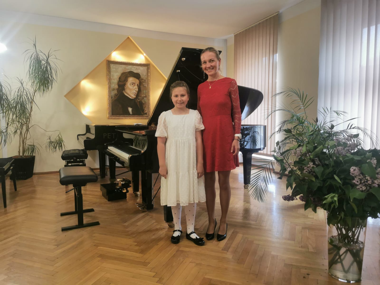 Na zdjęciu widoczna jest elegancko ubrana uczennica wraz z nauczycielką fortepianu, Panią Martą Łysyganicz. W tle widoczne są dwa fortepiany oraz obraz przedstawiający Fryderyka Chopina.