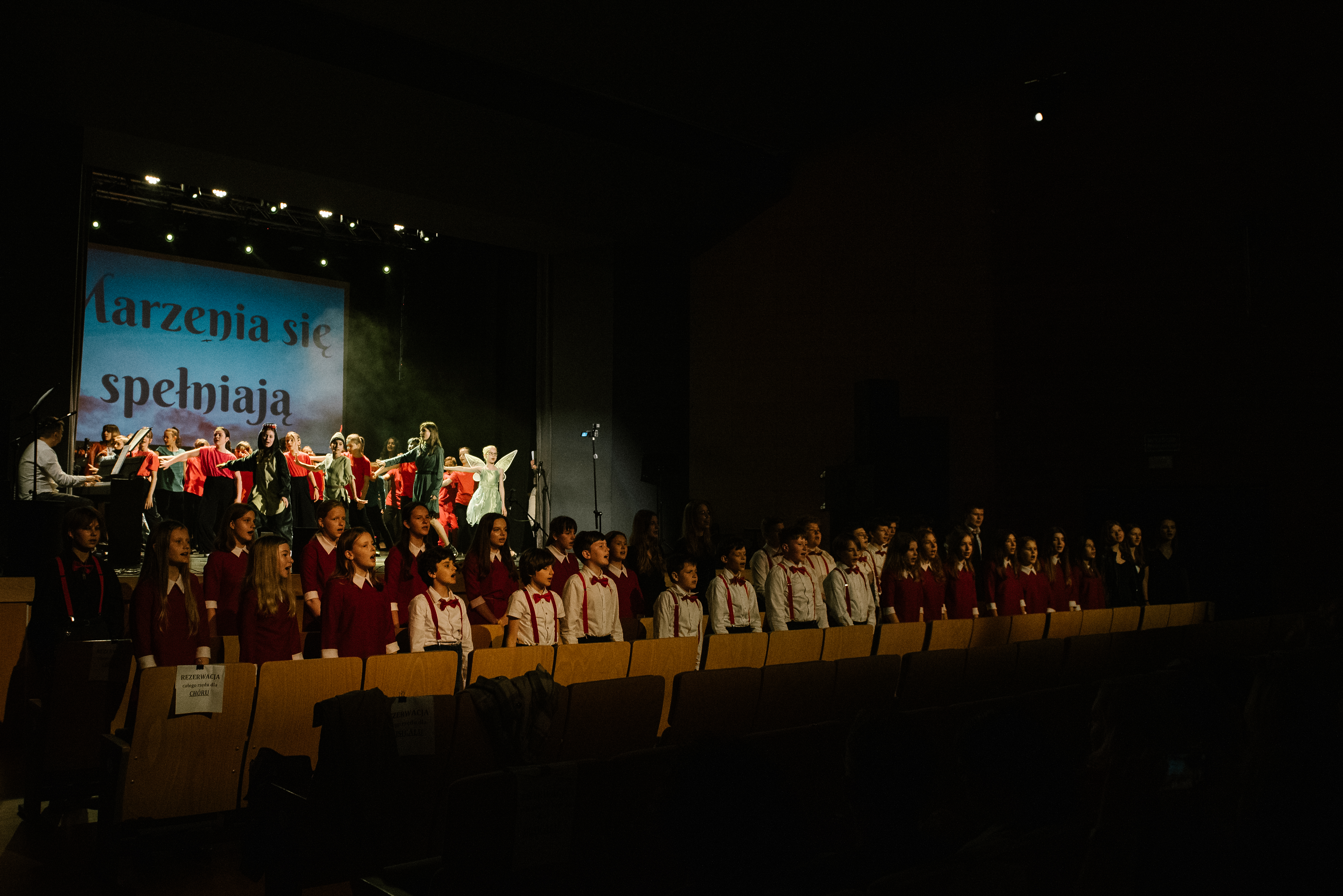 Na zdjęciu widoczna jest scena Strzelińskiego Ośrodka Kultury. W dwóch pierwszych rzędach widowni stoją dzieci należące do chóru szkolnego, natomiast na scenie znajdują się tańczący uczniowie w różnokolorowych strojach.