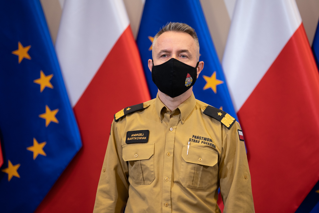 Zdjęcie przedstawia komendanta głównego Państwowej Straży Pożarnej Andrzeja Bartkowiaka na tle flag Polski i Unii Europejskiej.