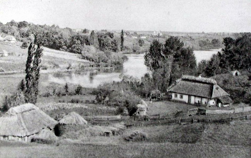 Czarno-białe zdjęcie przedstawiające pejzażowe ujęcie rodzinnego domu Szymanowskiego w Tymoszówce. Na pierwszym planie wiejski dom, zabudowania gospodarcze, drewniany płot, stogi siana. Za domem, płynąca rzeka. Dalej łagodne wzniesienie, pokryte w większej części liściastym lasem.