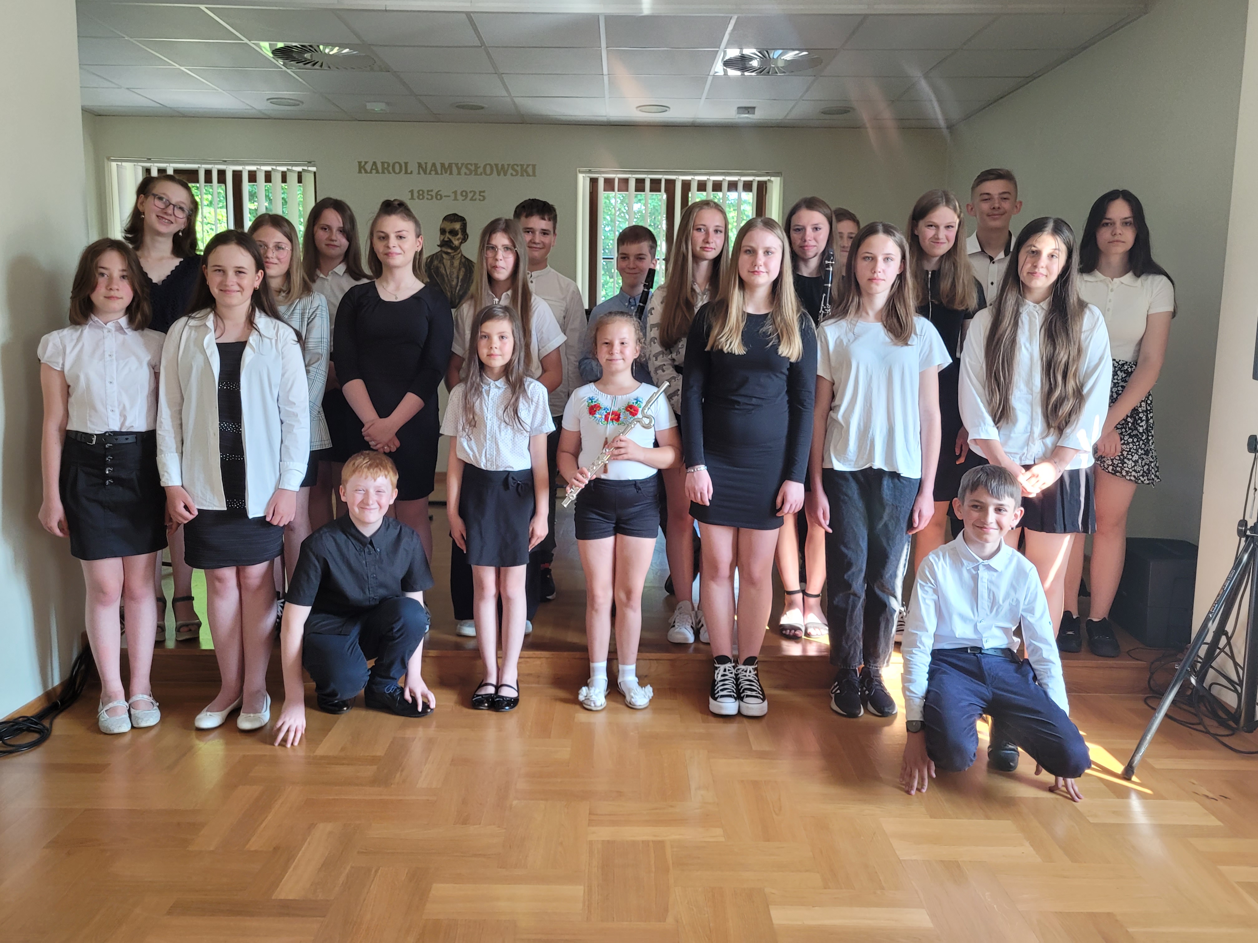 Sala koncertowa, przodem do publiczności ustawione dzieci -22osoby. Dzieci ubrane w stroje galowe. Dziewczynka stojąca pośrodku trzyma w dłoniach instrument -flet. Dwóch chłopców - jeden po lewej i jeden po prawej stronie kuca. W dali widać dwa okna oraz napis na ścianie Karol Namysłowski 1856-1925